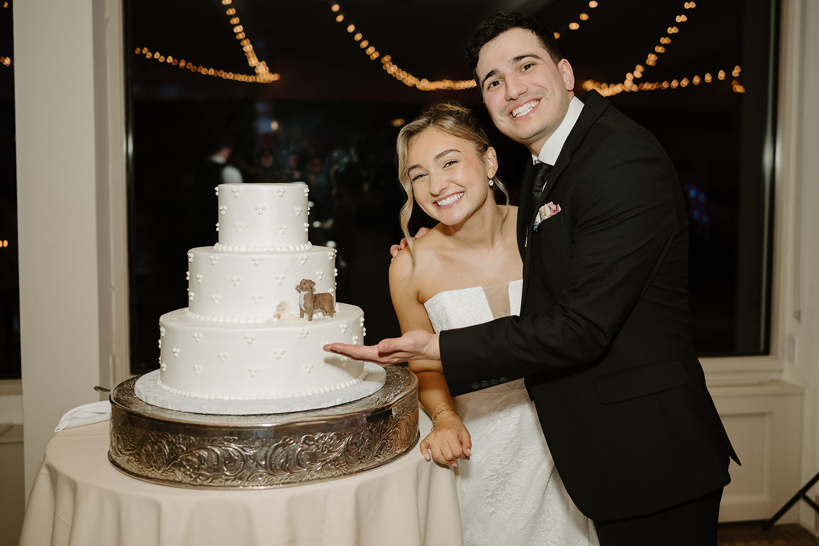 Newlyweds posing with their three-tier white wedding cake, pointing to a custom miniature dog figurine climbing up the side.