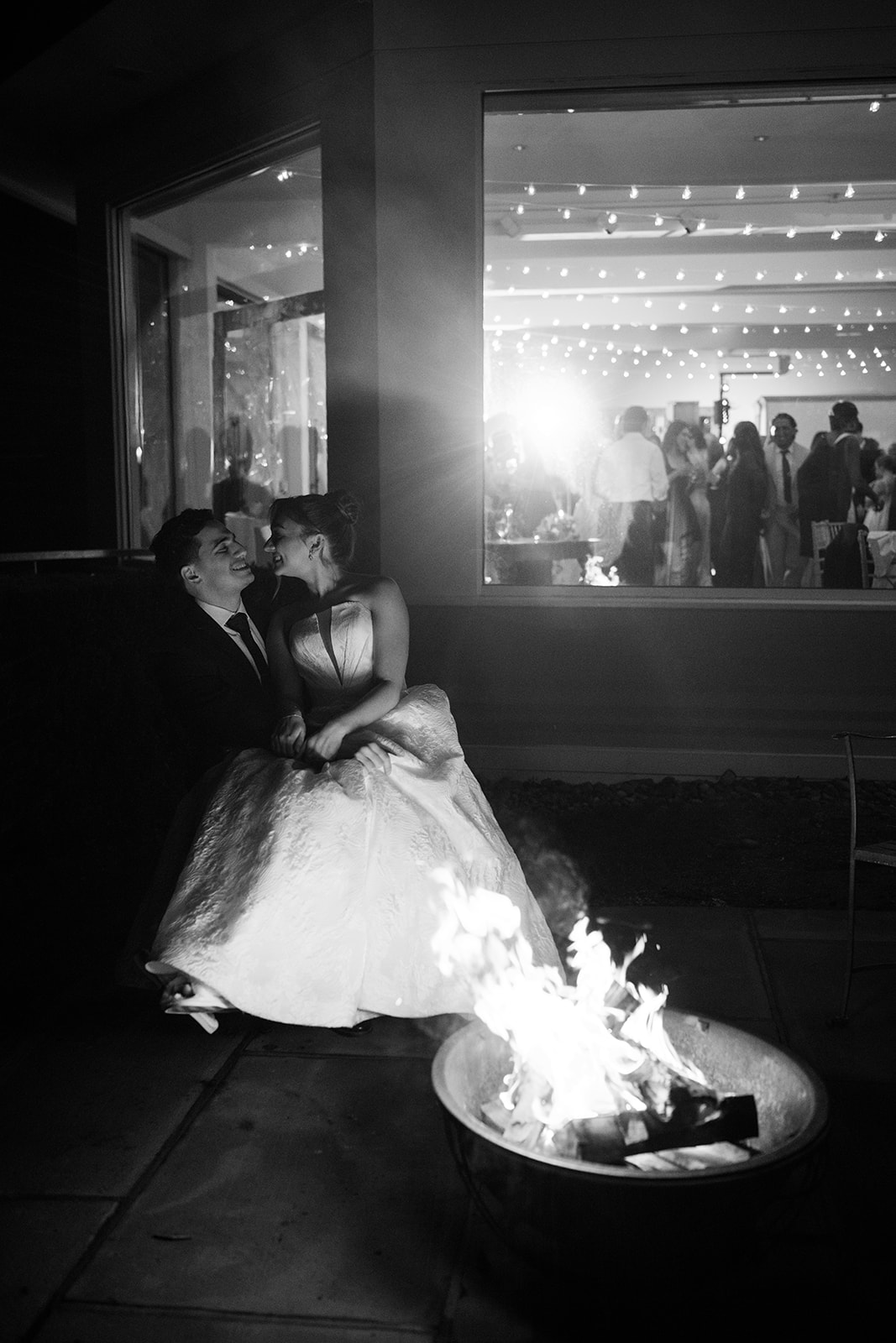 Black and white night photo of the bride and groom sitting outside by a glowing bonfire fire pit, smiling at each other.
