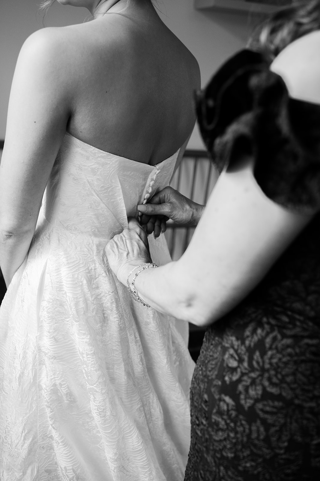 Close-up B&W shot of the mother buttoning the delicate button details on the back of the bride's wedding gown.
