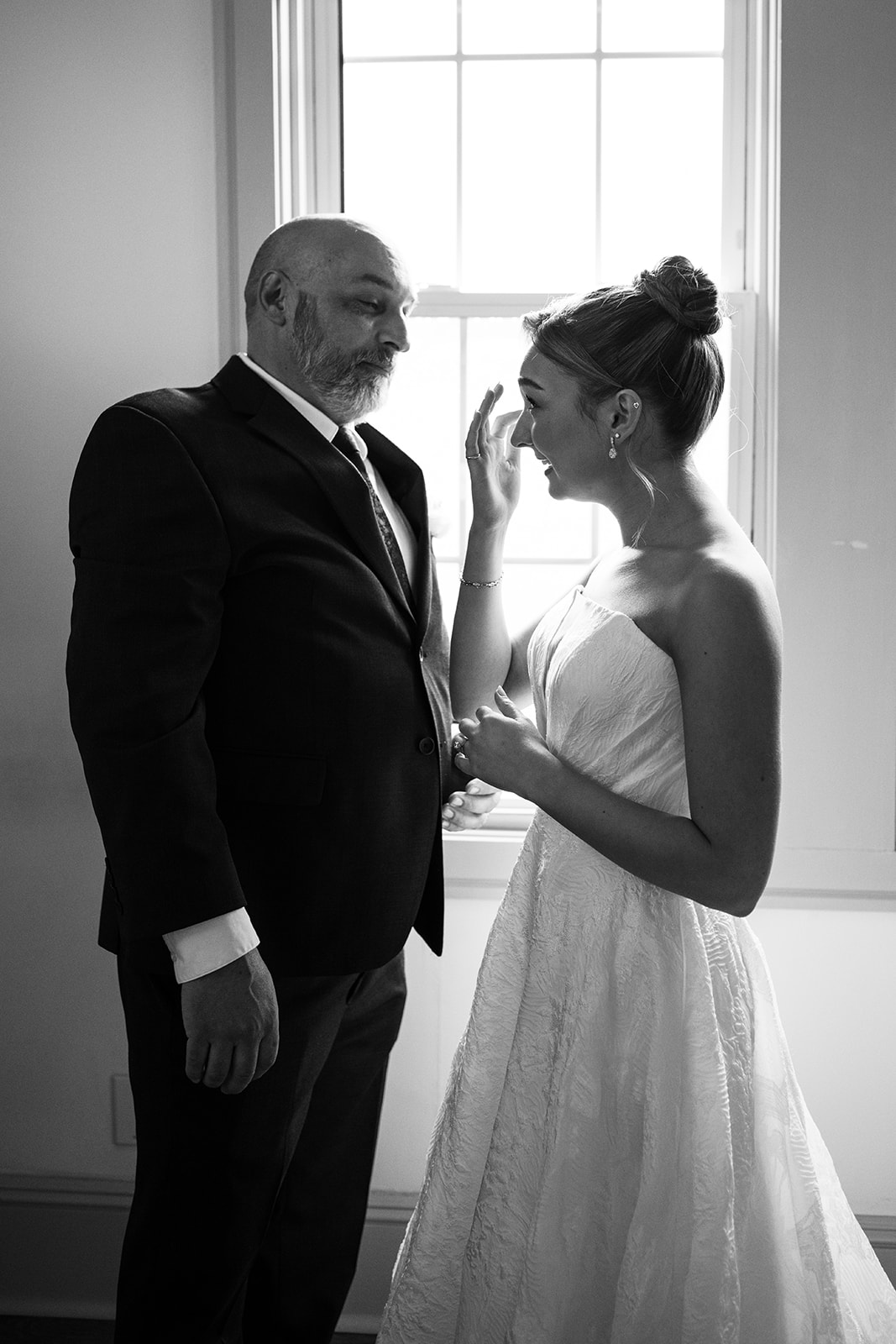 Black and white photo of the bride wiping away a tear while her father looks at her emotionally during their first look in front of a window.