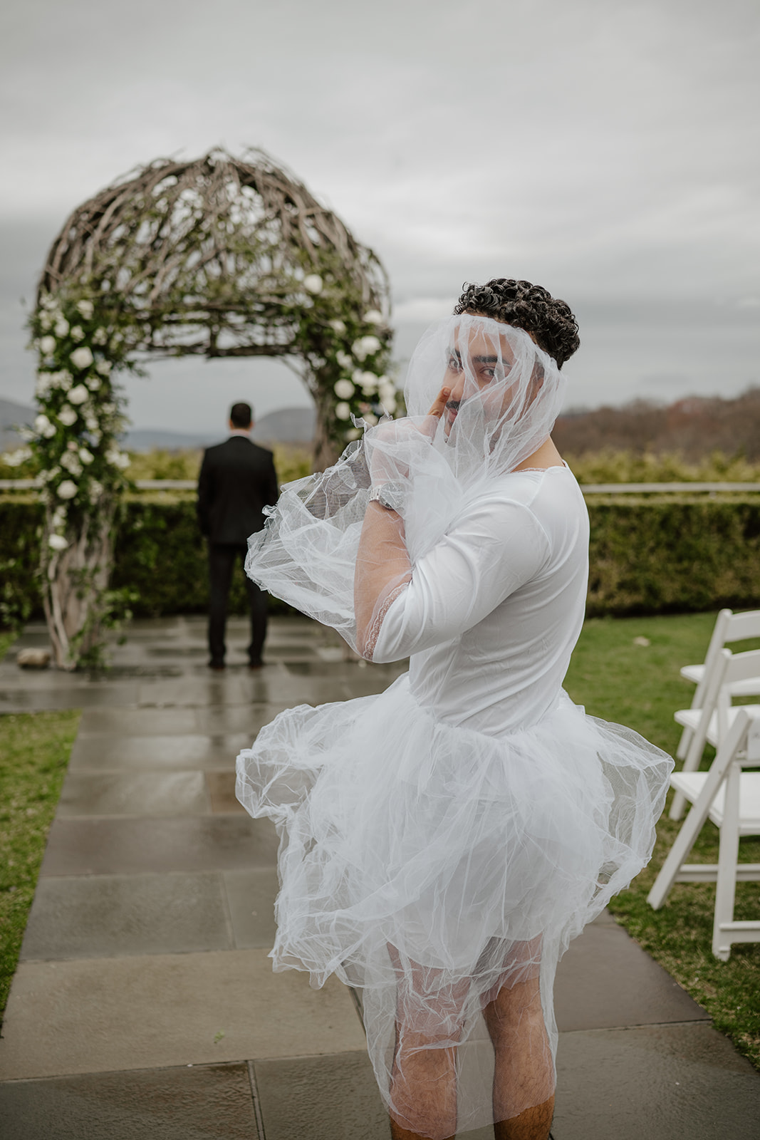 Groomsman/best man dressed in a white dress/tutu and a wedding veil over his face, holding a finger to his lips (shushing), walking up to the groom who has his back turned waiting for a first look under a wooden garden arch.
