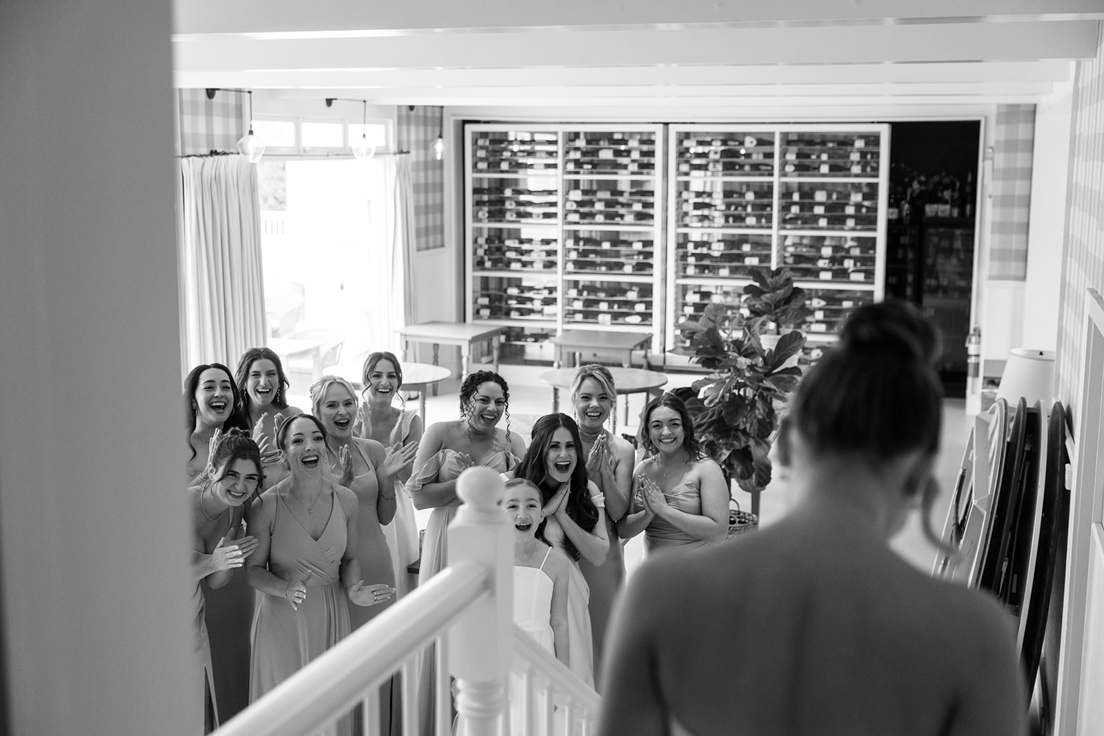 Black and white photo showing the bridesmaids gathered at the bottom of the stairs, looking up at the bride with expressions of joy and surprise.