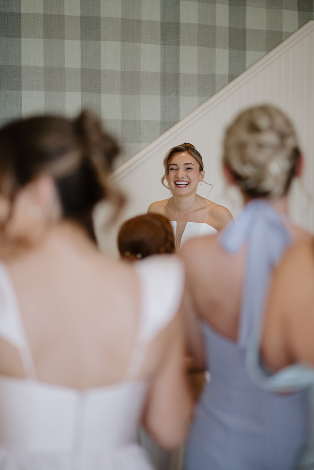 Color photo showing the bride smiling and laughing in the center, framed by the out-of-focus backs of her bridesmaids looking at her.