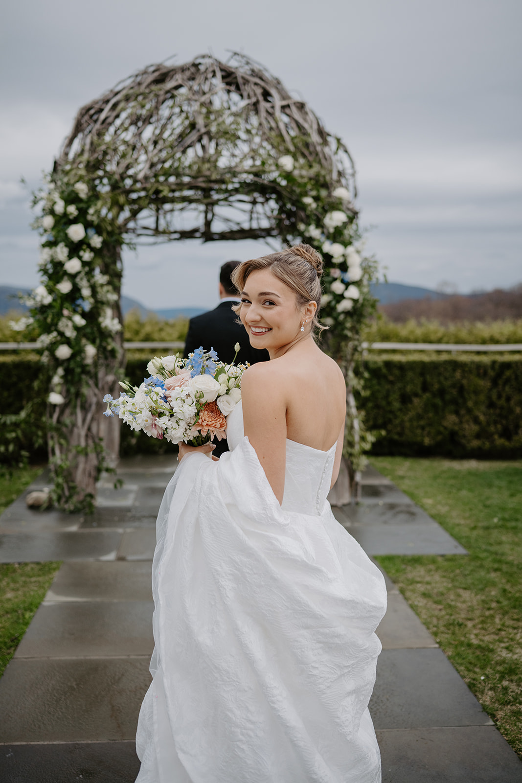 Bride smiling and looking over her shoulder at the camera, holding her bouquet, as she walks up to the groom who has his back turned waiting for the real first look under the wooden garden arch.