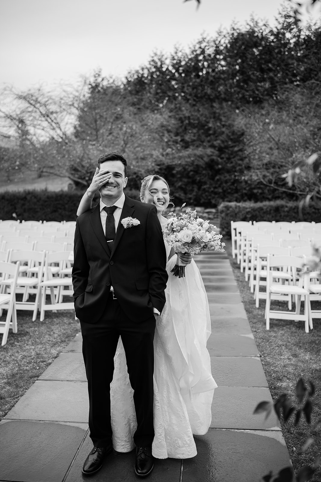 Black and white photo of the bride walking up behind the groom and covering his eyes with her hand, both smiling, with rows of white ceremony chairs on either side.