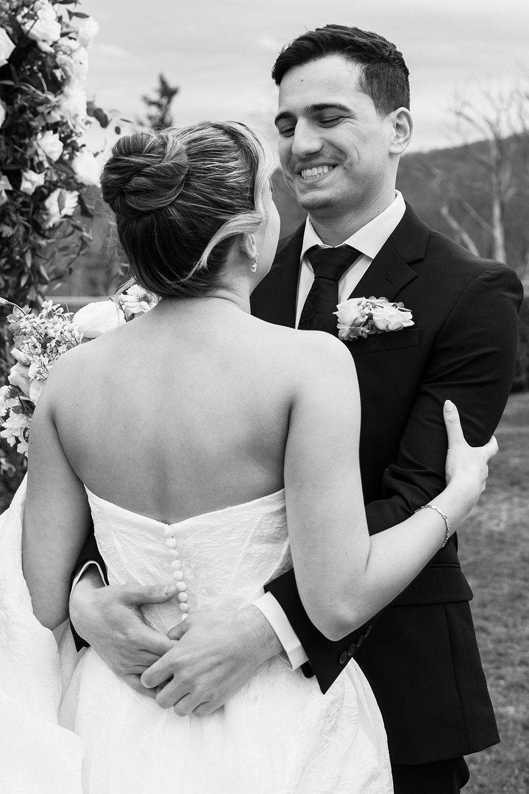 Black and white photo of the groom hugging the bride from behind/holding her waist, while she looks over her shoulder towards him, showing the back details of her strapless wedding dress with delicate buttons.