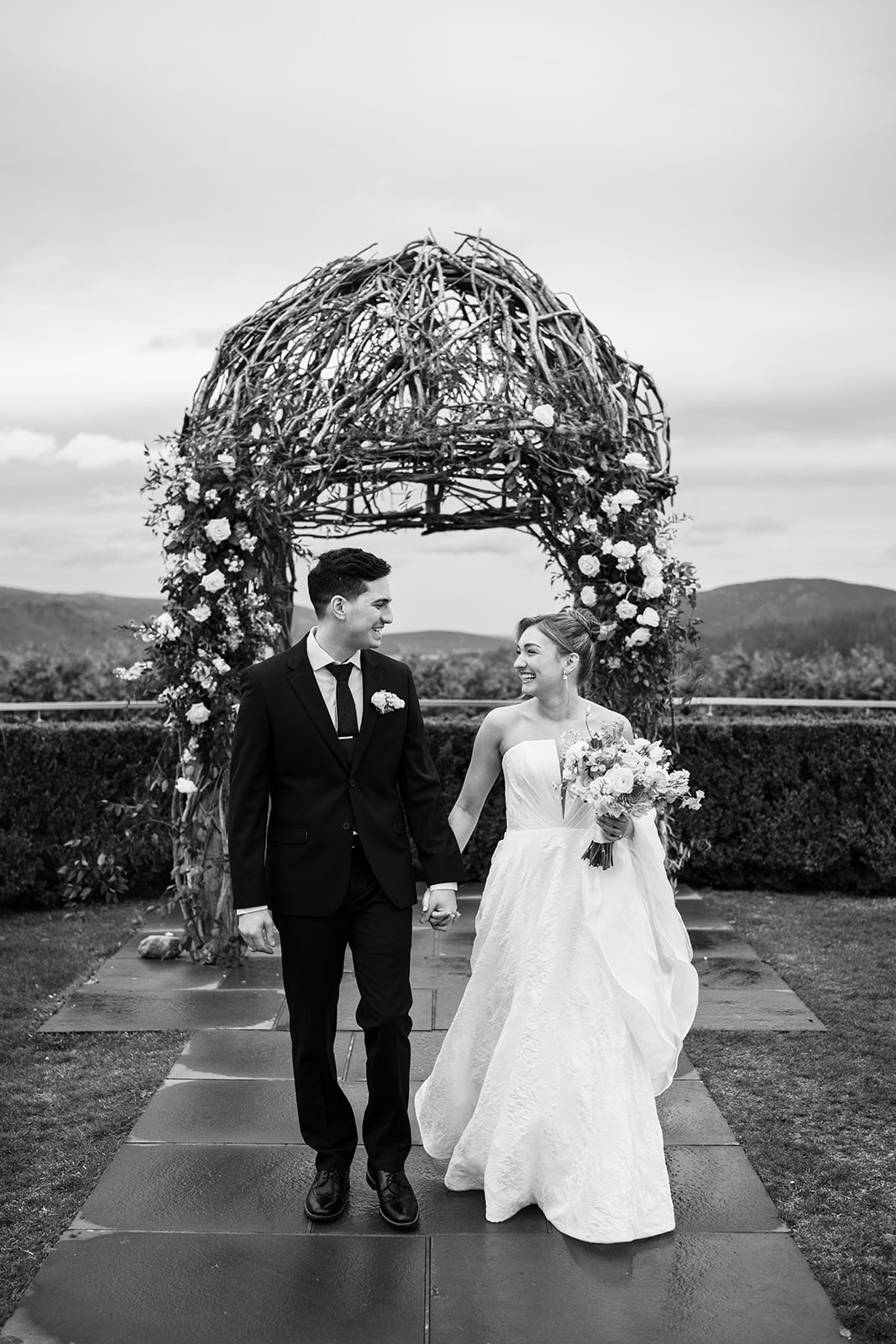 Black and white photo of the bride and groom holding hands, smiling and walking away from the wooden ceremony arch.