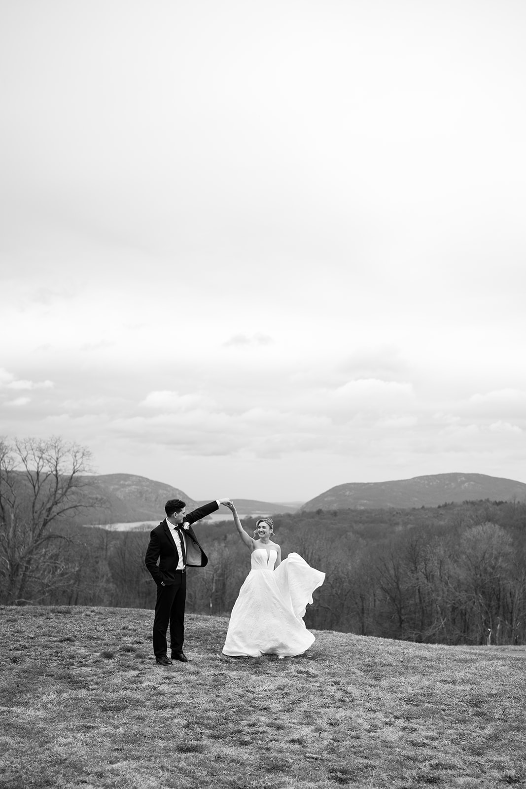 Black and white photo of the groom twirling the bride on a grassy hill overlooking the valley.