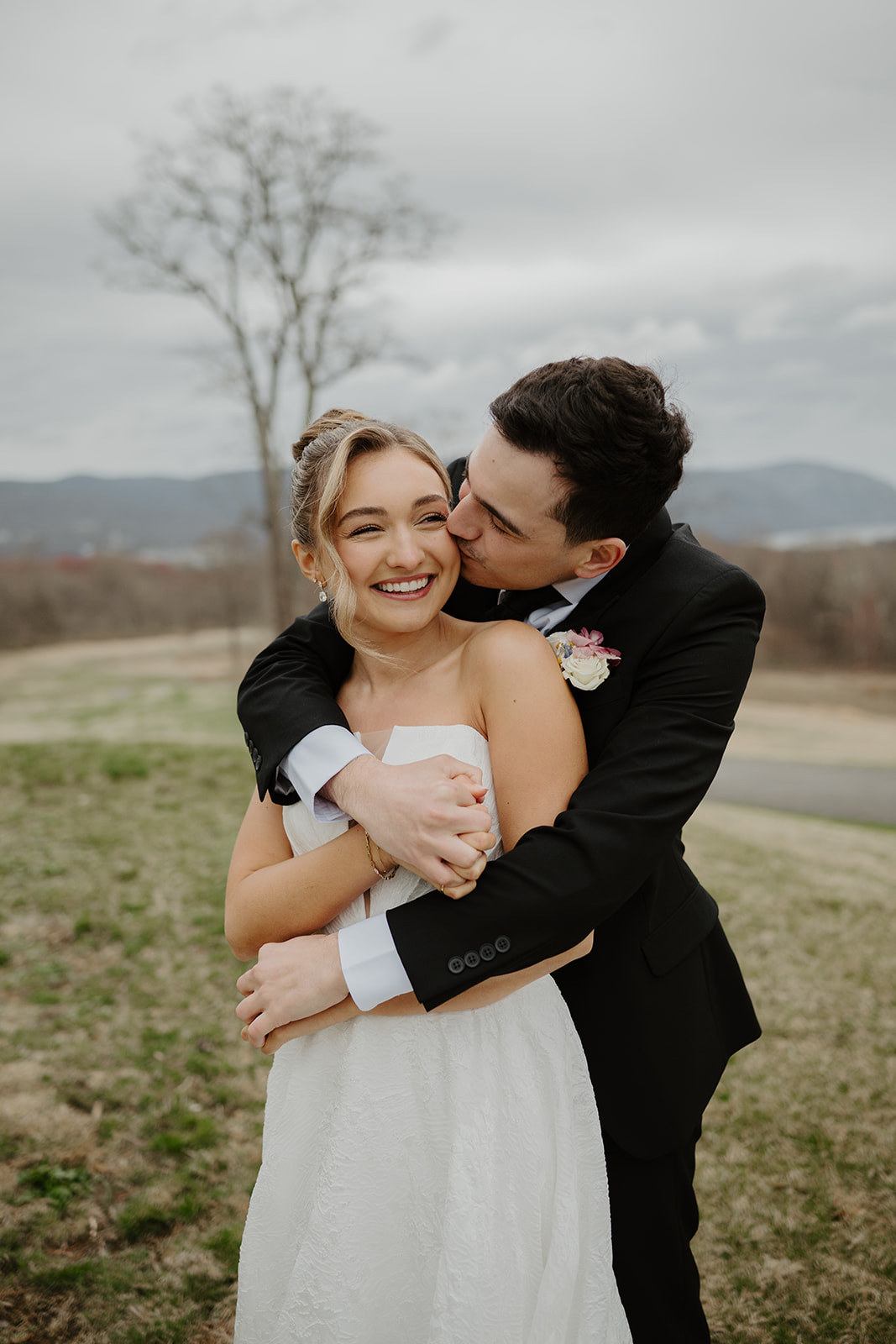 Color photo of the groom hugging the bride from behind, kissing her cheek on the grassy overlook.