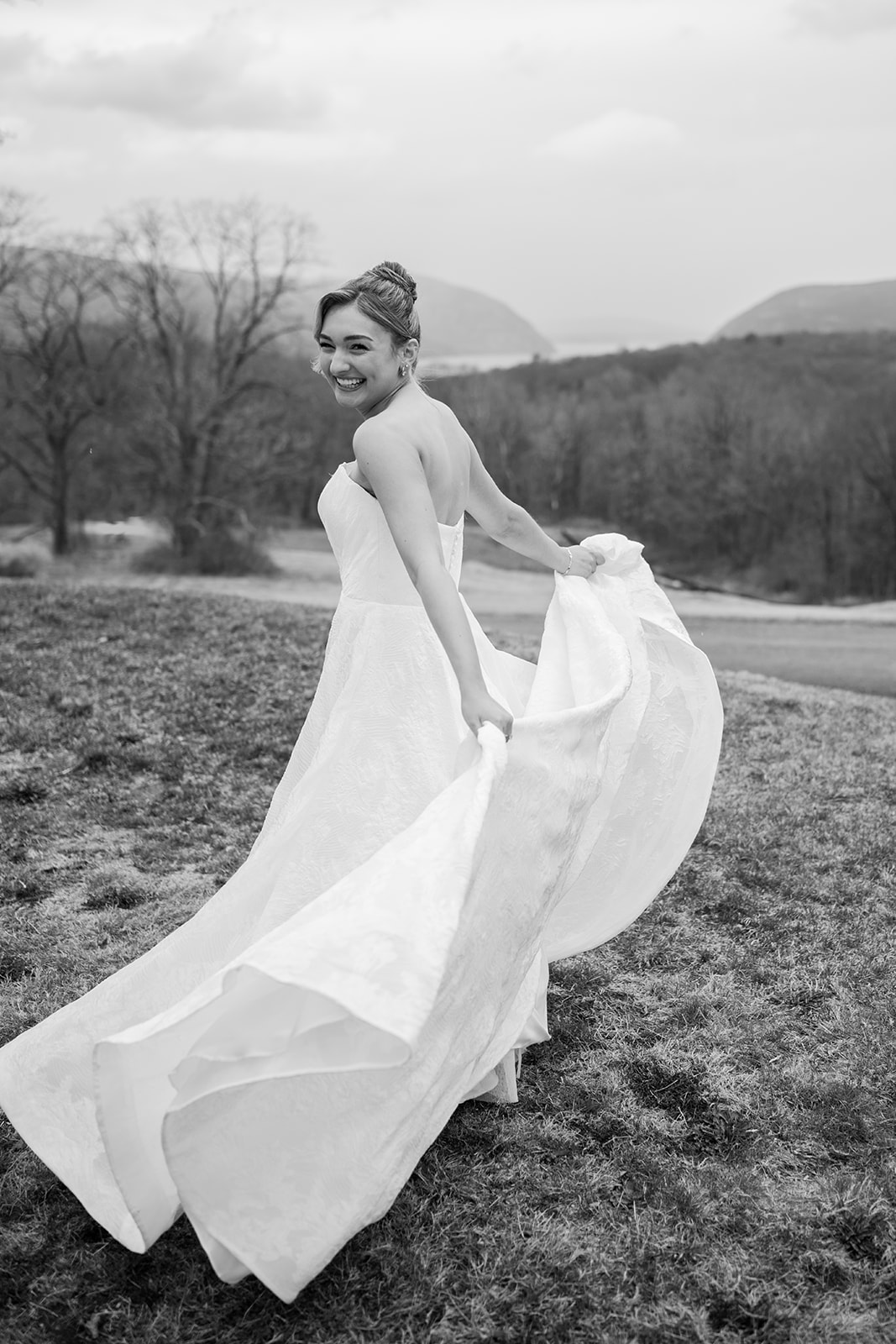 Black and white photo of the bride walking through fields carrying her wedding dress train.