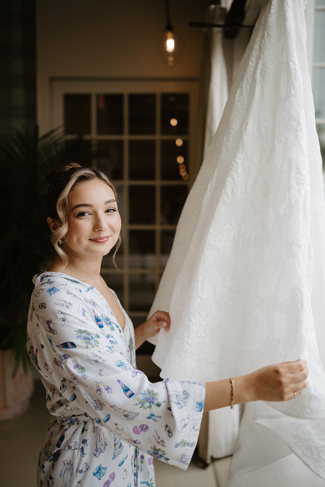 Bride in patterned silk robe looking at camera and smiling with one hand on her wedding gown hanging in front of a window.
