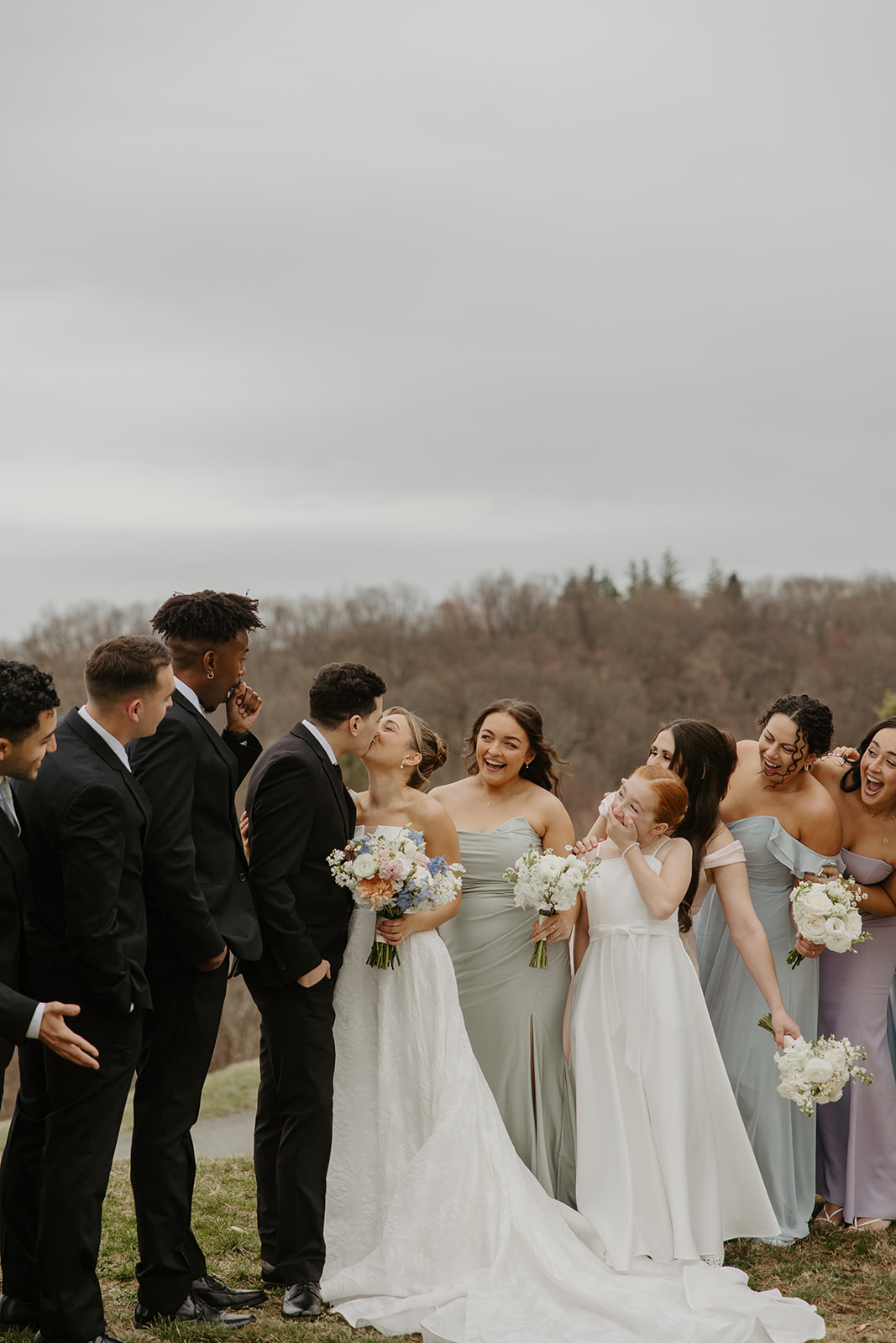 Wedding party laughing as the couple shares a kiss on the grassy lawn.