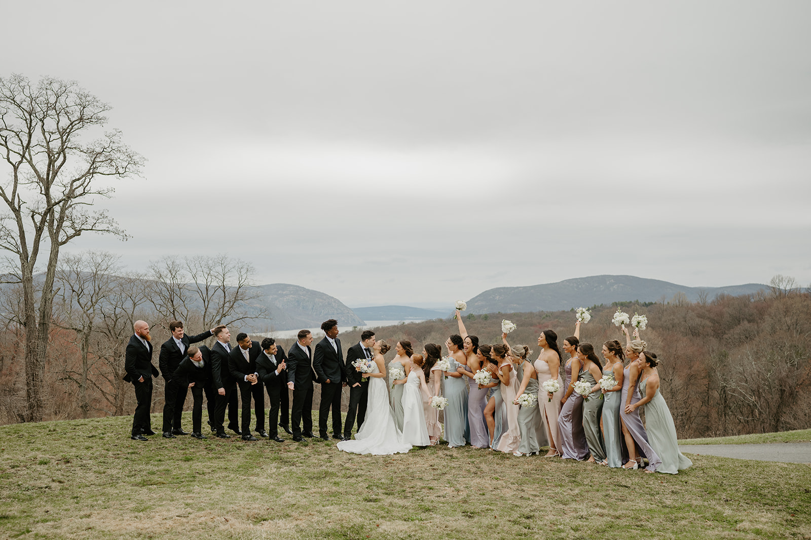 Full wedding party standing in a long line on a hill, cheering as the couple kisses.