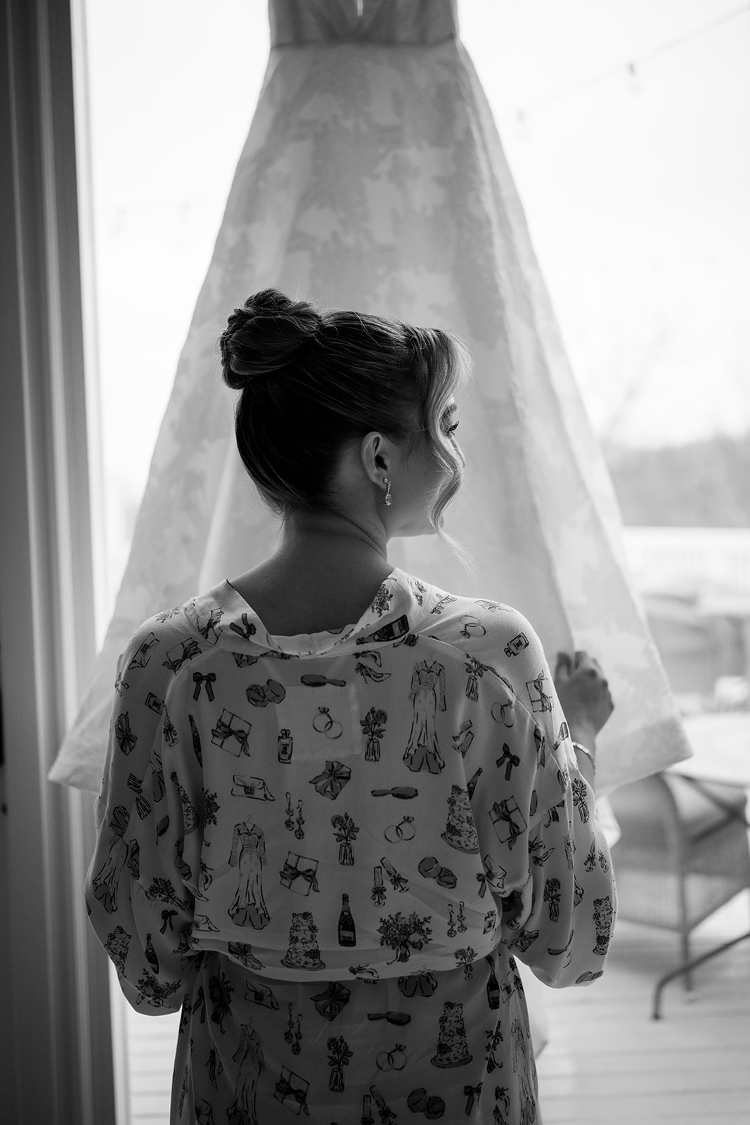 Black and white photo of the bride from behind in a patterned silk robe looking towards her wedding gown hanging in the window.