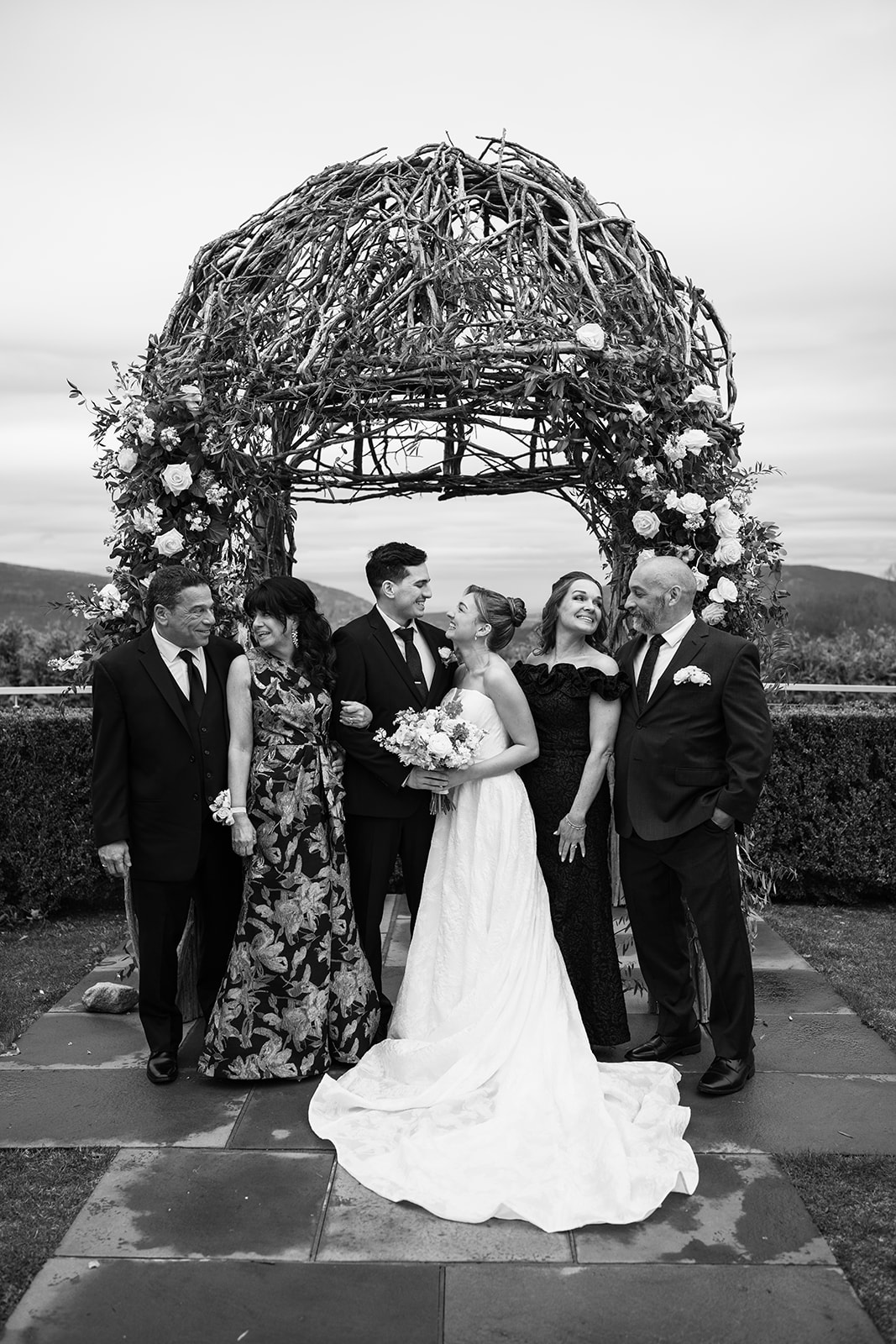 Black and white photo of the couple posing with their parents under the ceremony arch.