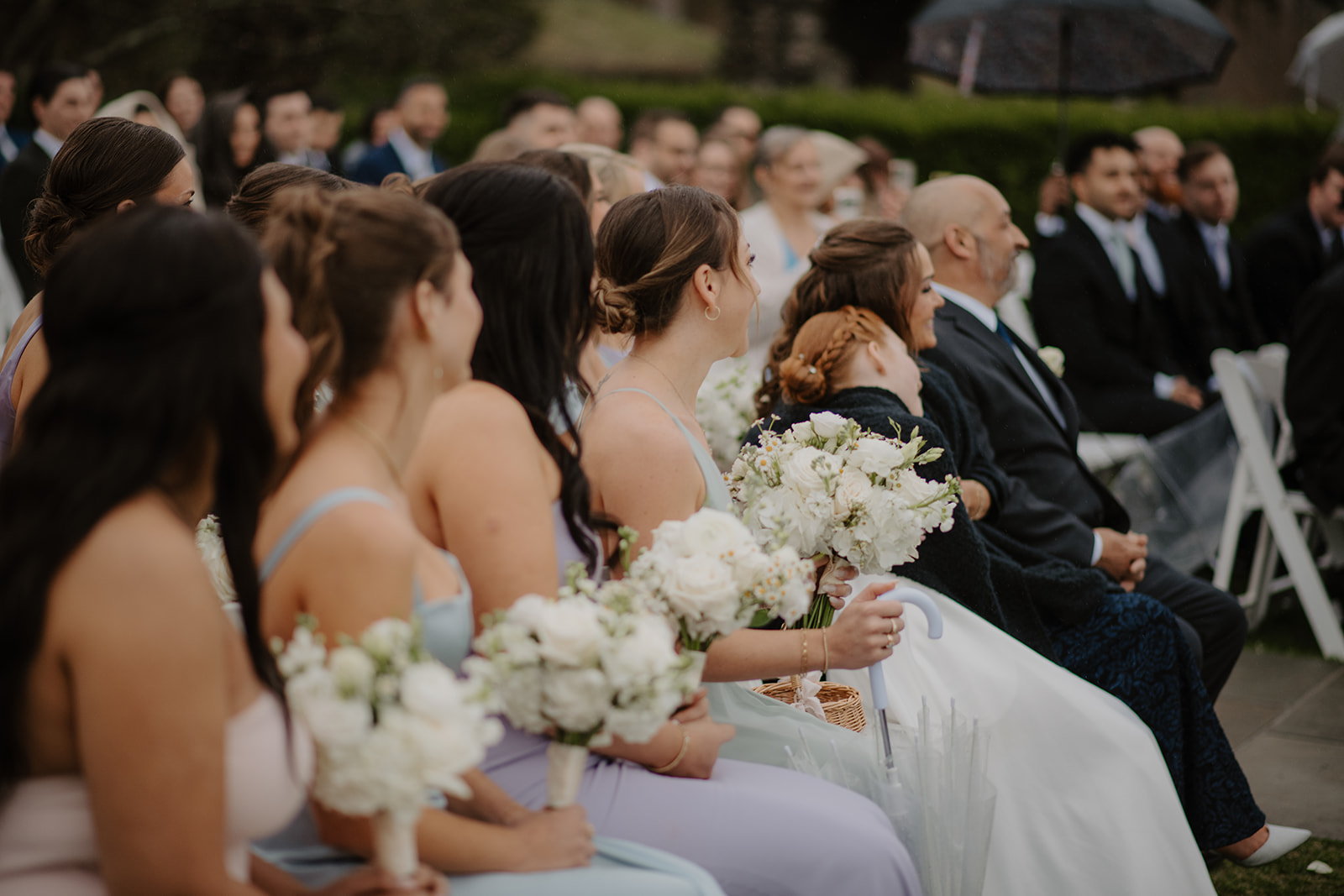 Bridesmaids and guests seated in rows under clear umbrellas, watching the ceremony.