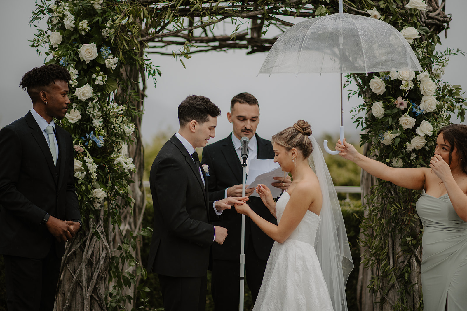 Exchange of wedding rings under the arch, with a bridesmaid holding a clear umbrella over the bride.