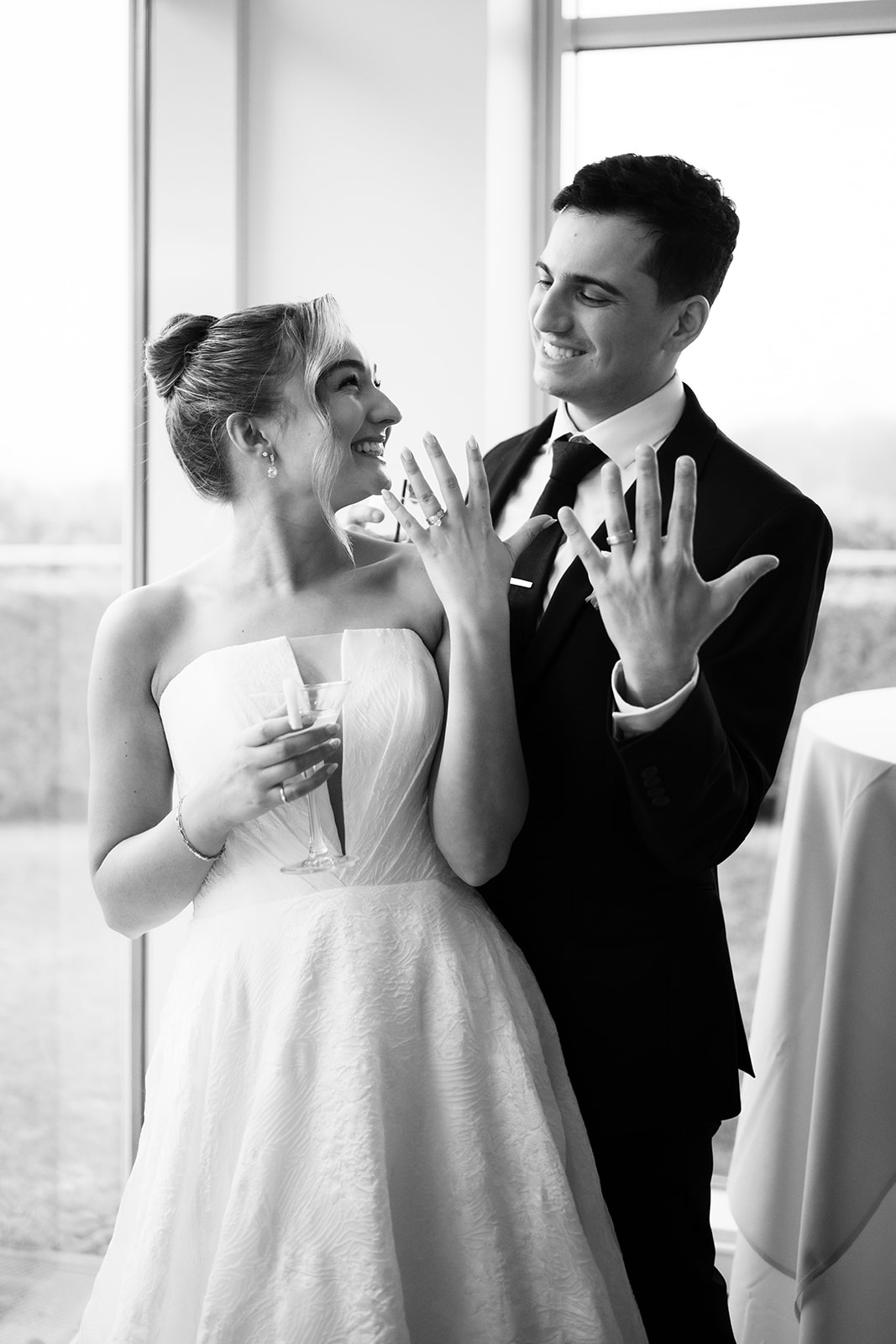Black and white photo of the couple showing off their wedding rings, the bride holding a cocktail.
