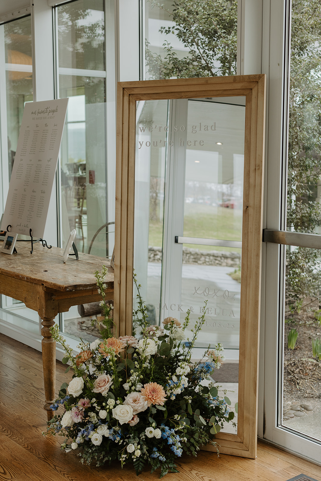 Wooden-framed welcome mirror sign reading 'we're so glad you're here / xoxo, / JACK & BELLA / 04.05.25' decorated with a floral arrangement at the base.