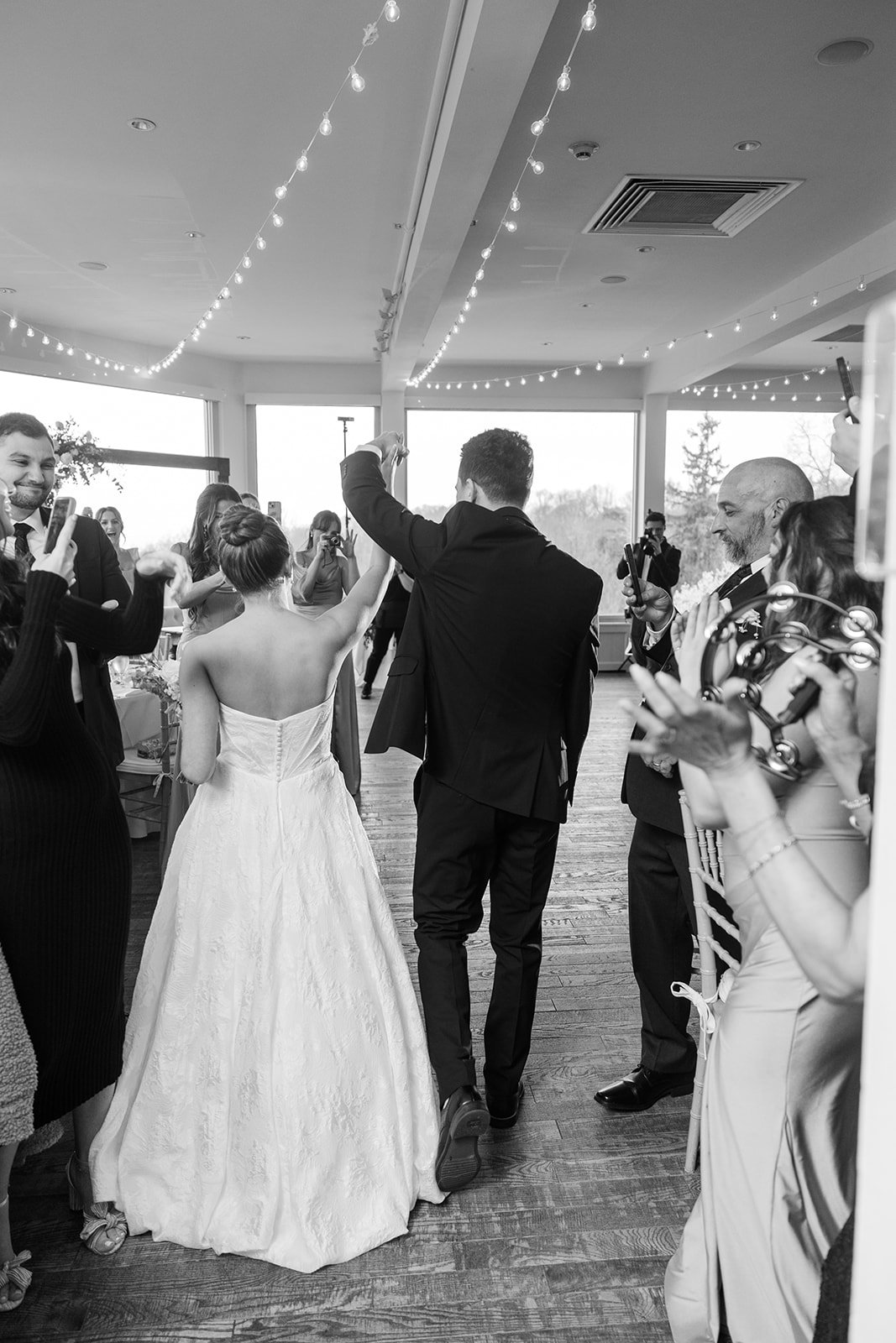 Black and white photo of the couple's grand entrance into the reception ballroom from behind, holding hands high while guests cheer.