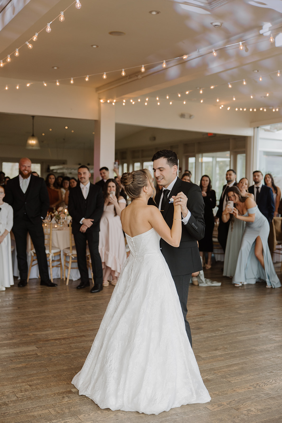 Newlyweds holding hands and sharing their romantic first dance inside the reception ballroom at The Garrison under warm string lights.