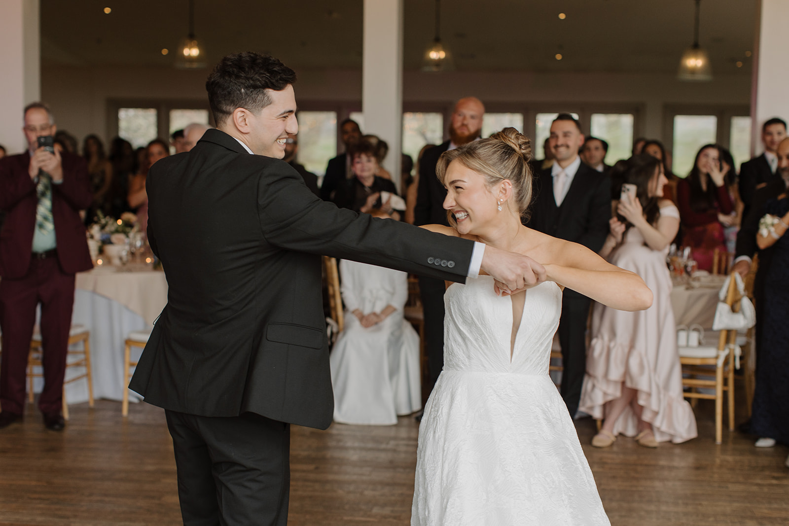 Groom smiling as he twirls the bride during their first dance, with wedding guests watching in the background.