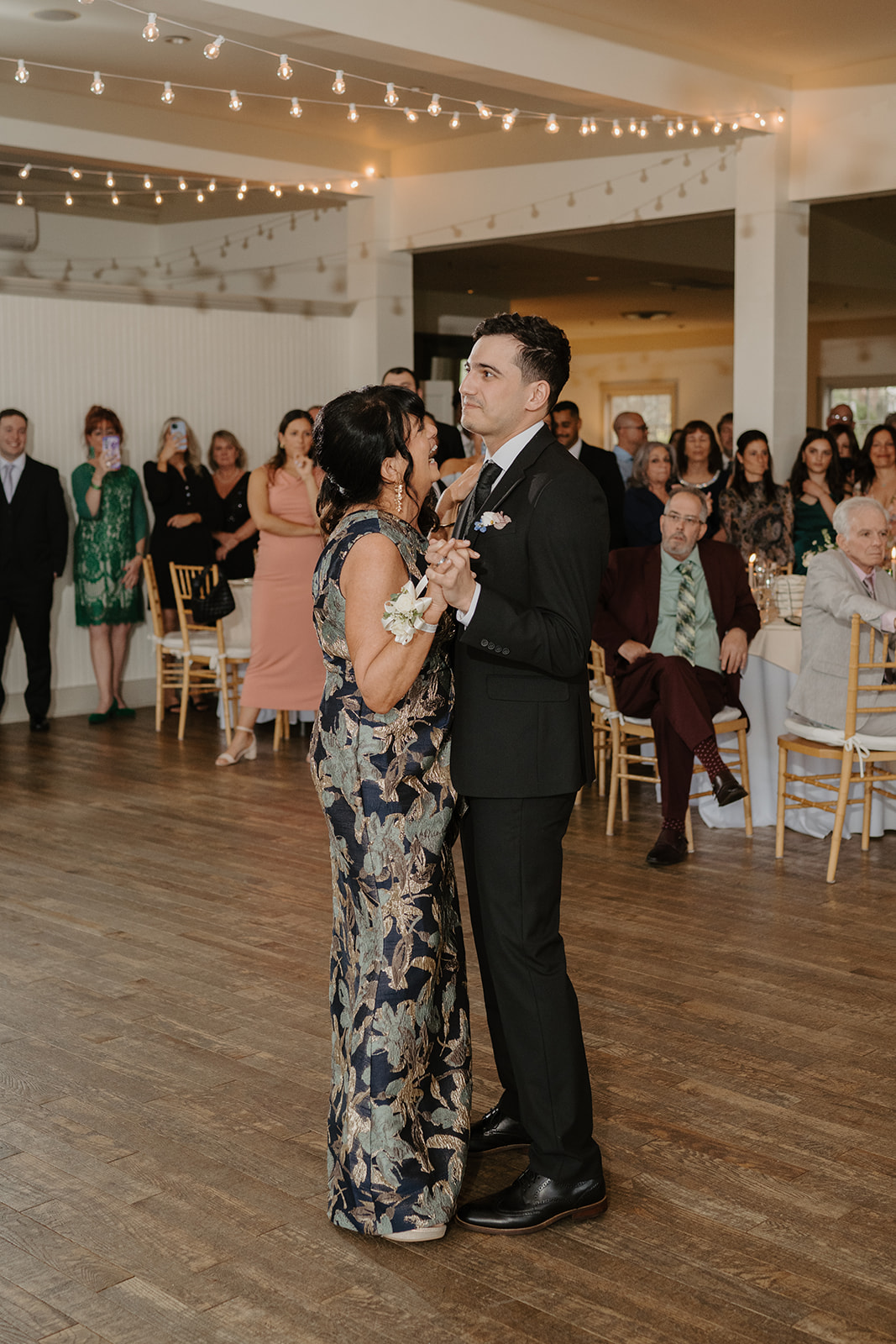 Groom and his mother sharing a sweet dance at the reception, both smiling, surrounded by seated wedding guests.