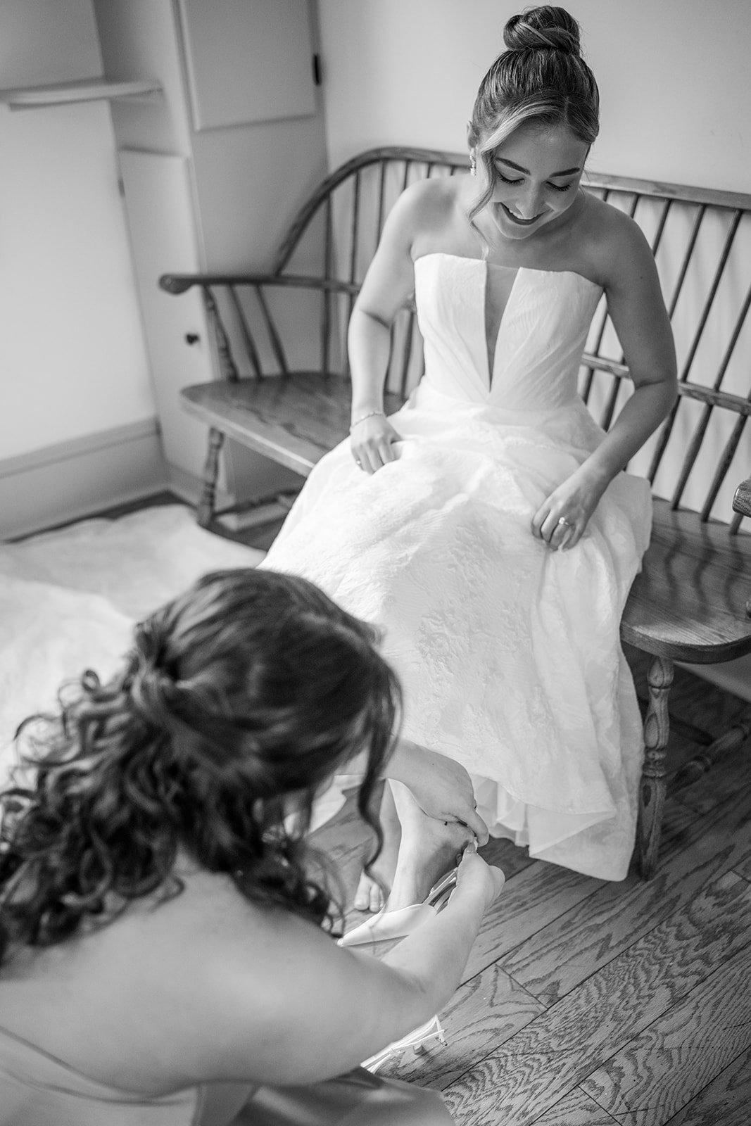 Black and white photo of the bride sitting on a wooden bench, smiling, as her bridesmaid helps her put on her white wedding heels.