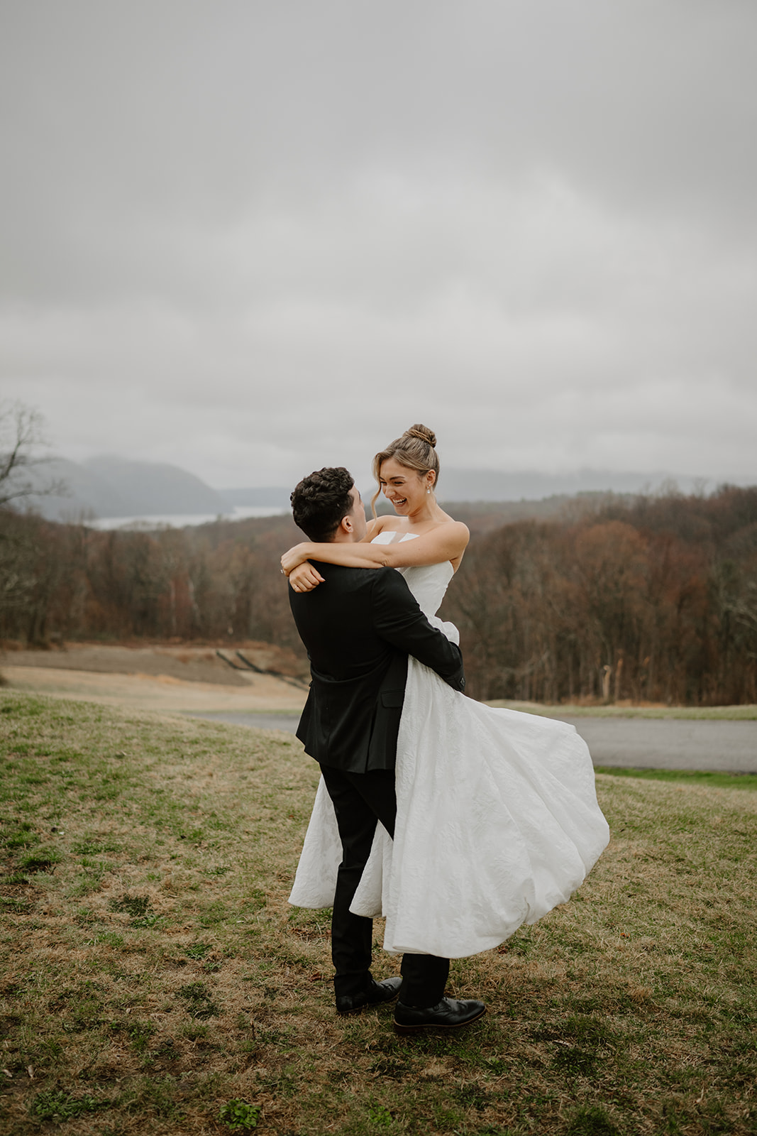 Groom lifting and spinning the laughing bride on the grassy overlook at The Garrison, with the Hudson Highlands mountains in the background.