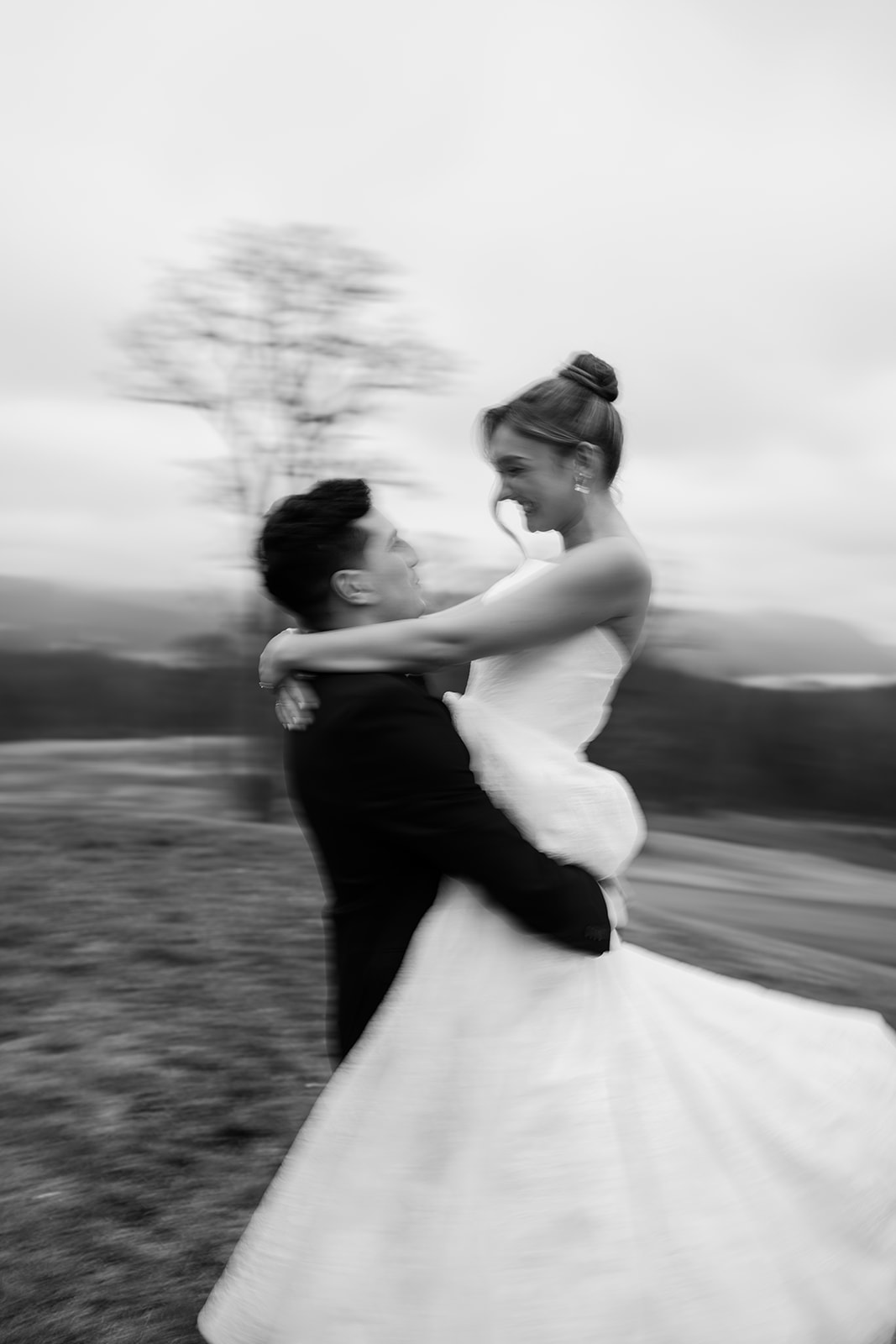 Artistic black and white photo with motion blur showing the groom lifting and spinning the bride on the hillside at dusk.