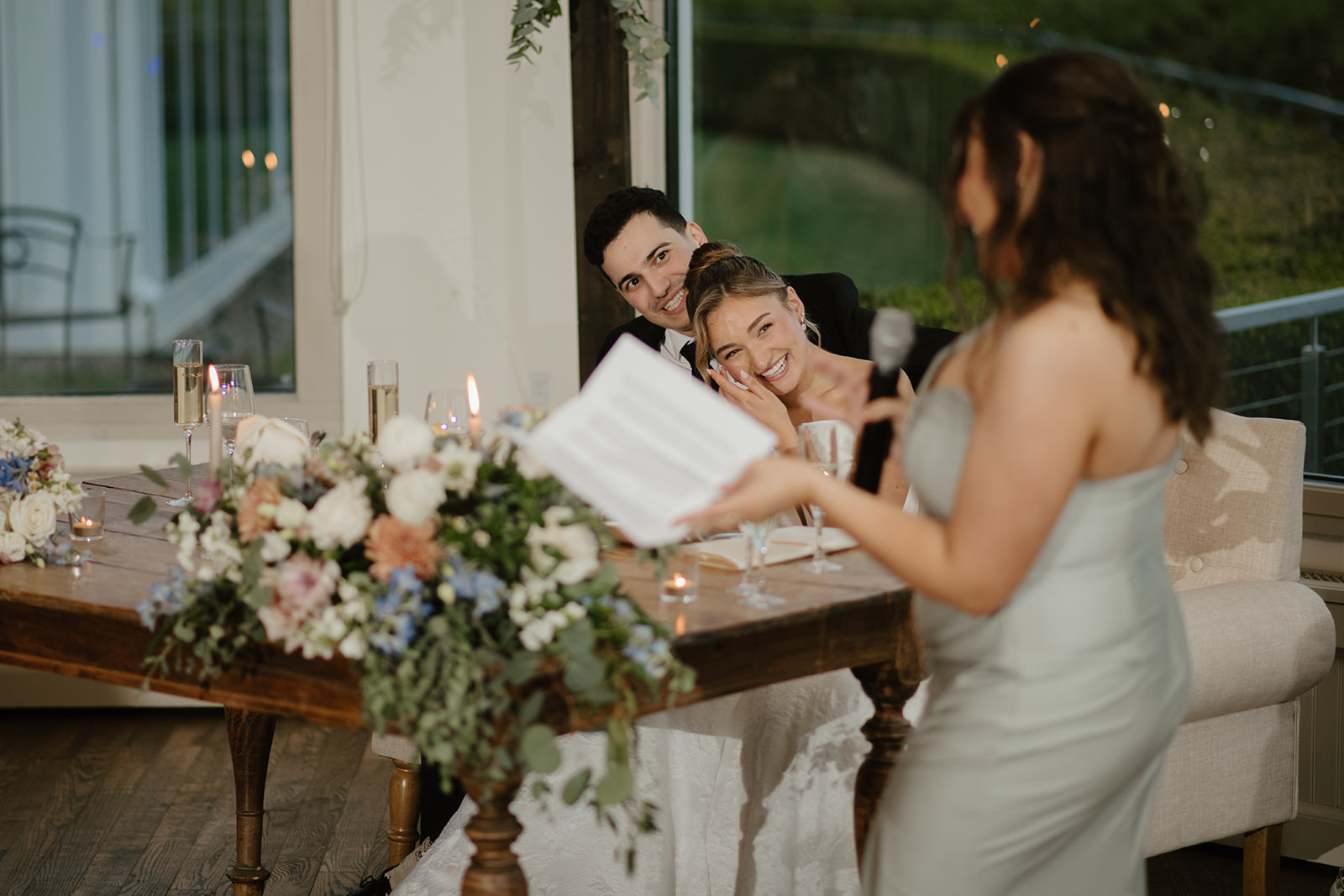 Maid of honor delivering a heartfelt toast, while the bride and groom laugh and smile at their sweetheart table.