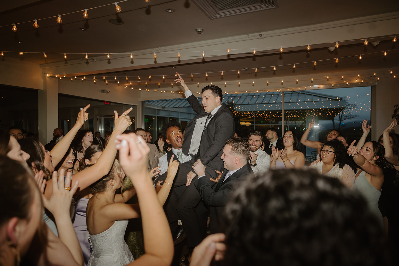 Groom celebrating on the shoulders of his friends, waving one hand in the air on the crowded dance floor.