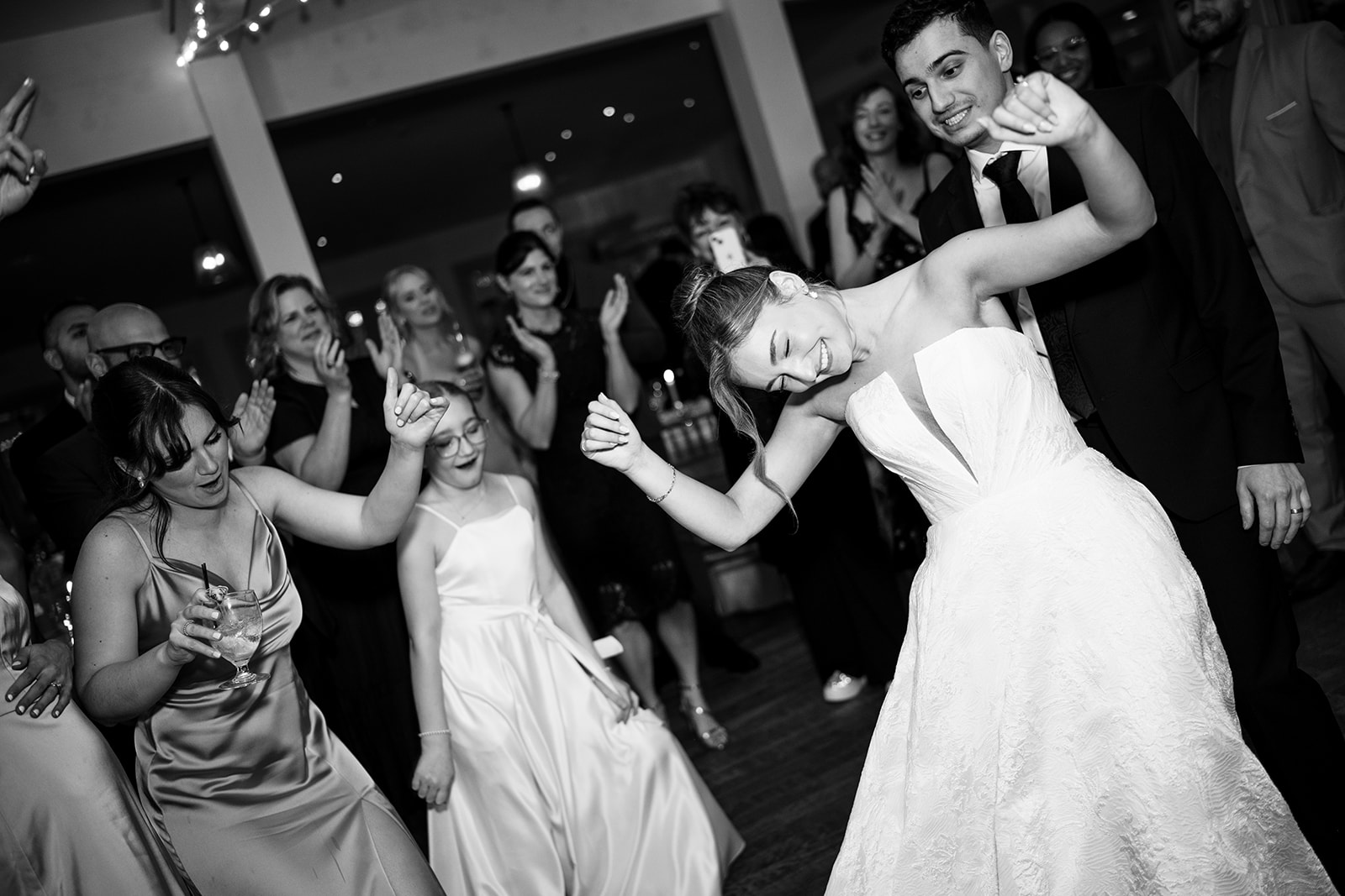 Black and white photo of the bride dancing enthusiastically in the center of a circle of cheering wedding guests.
