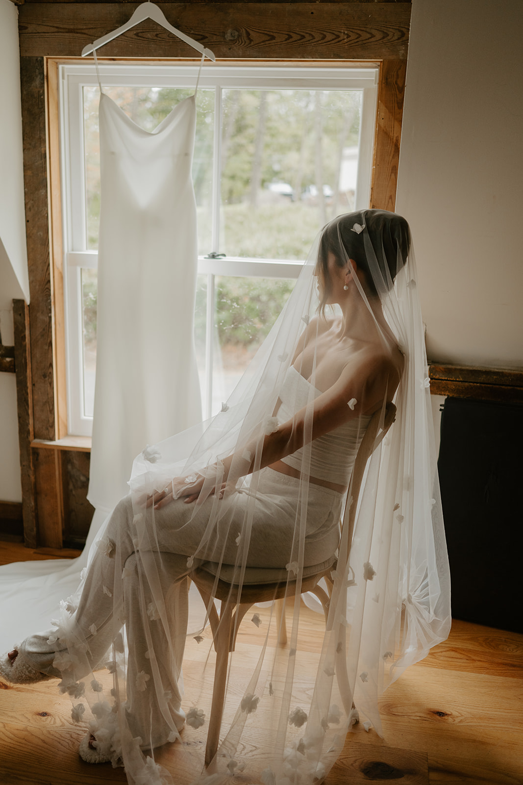Bride sits draped in a delicate floral veil next to her wedding gown hanging in the window.