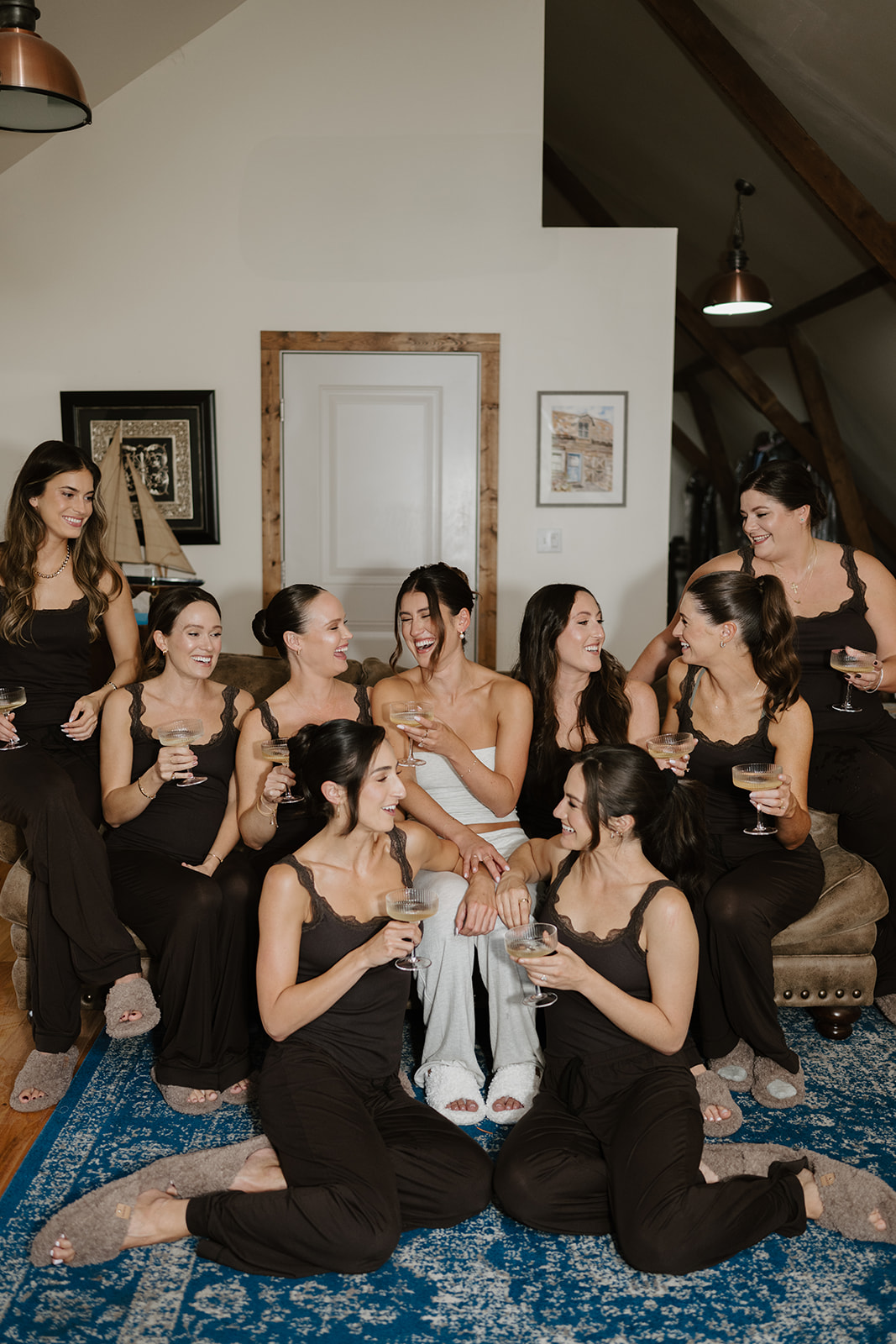 Bride and bridesmaids laugh while holding champagne glasses on a couch in the bridal suite.