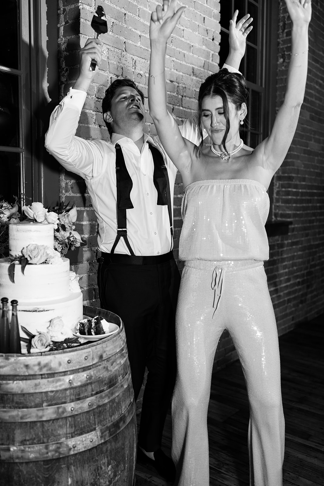 Black and white photo of the bride and groom dancing joyfully by their wedding cake.