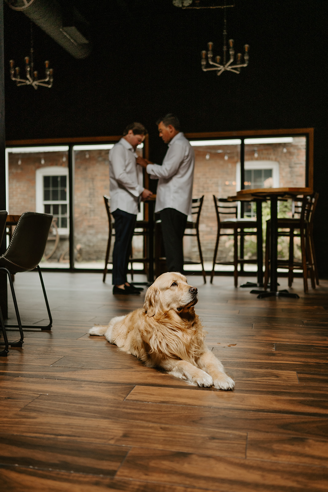 Golden retriever rests on the wooden floor as the groom and best man prepare in the background.