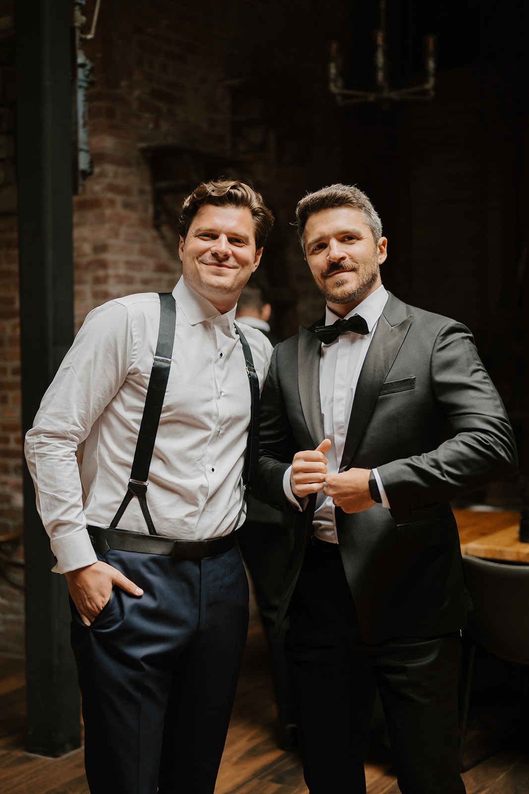 Groom in suspenders smiles beside his best man in a tuxedo inside a brick venue.