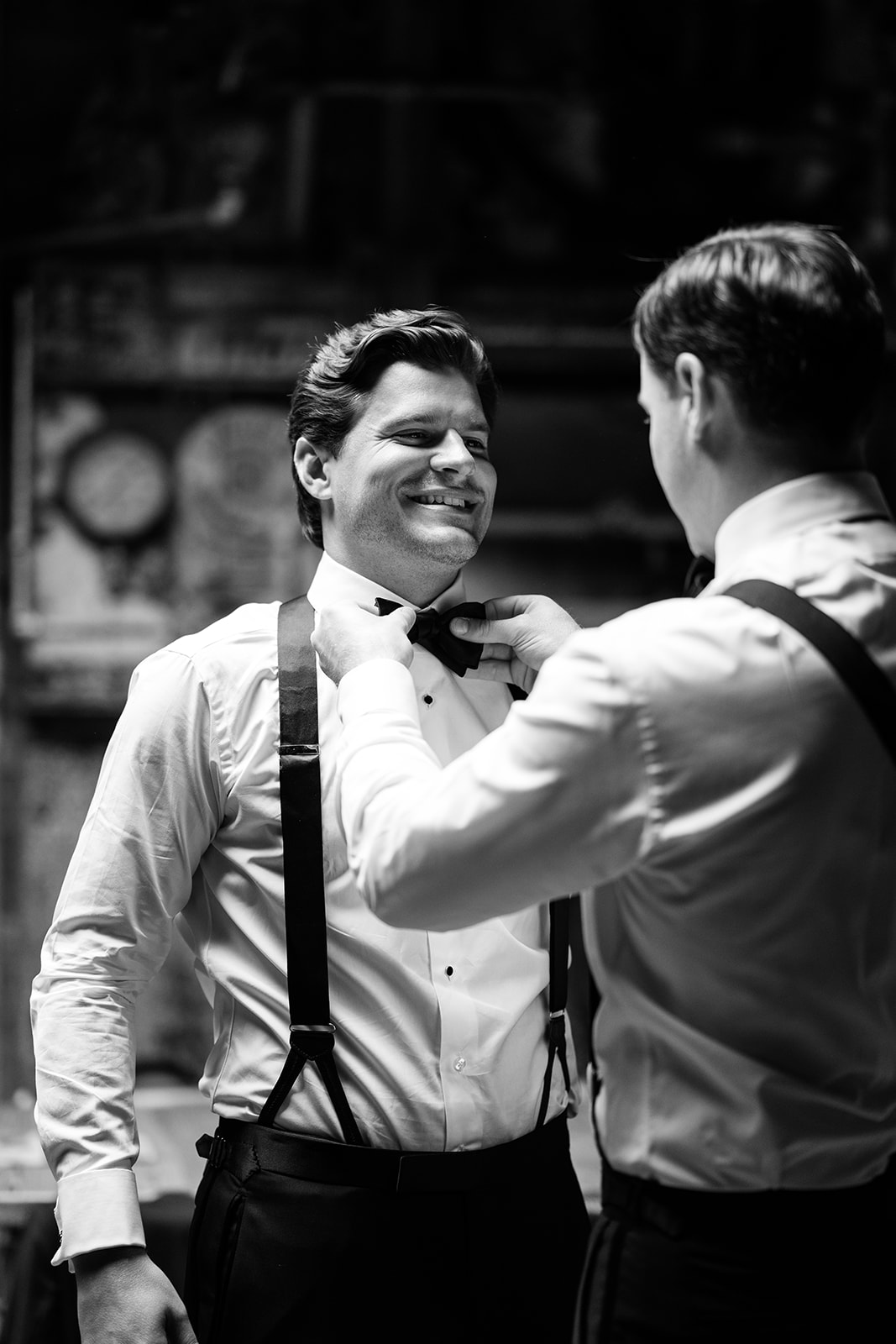 Black and white portrait of the groom smiling as his bow tie is adjusted before the ceremony.