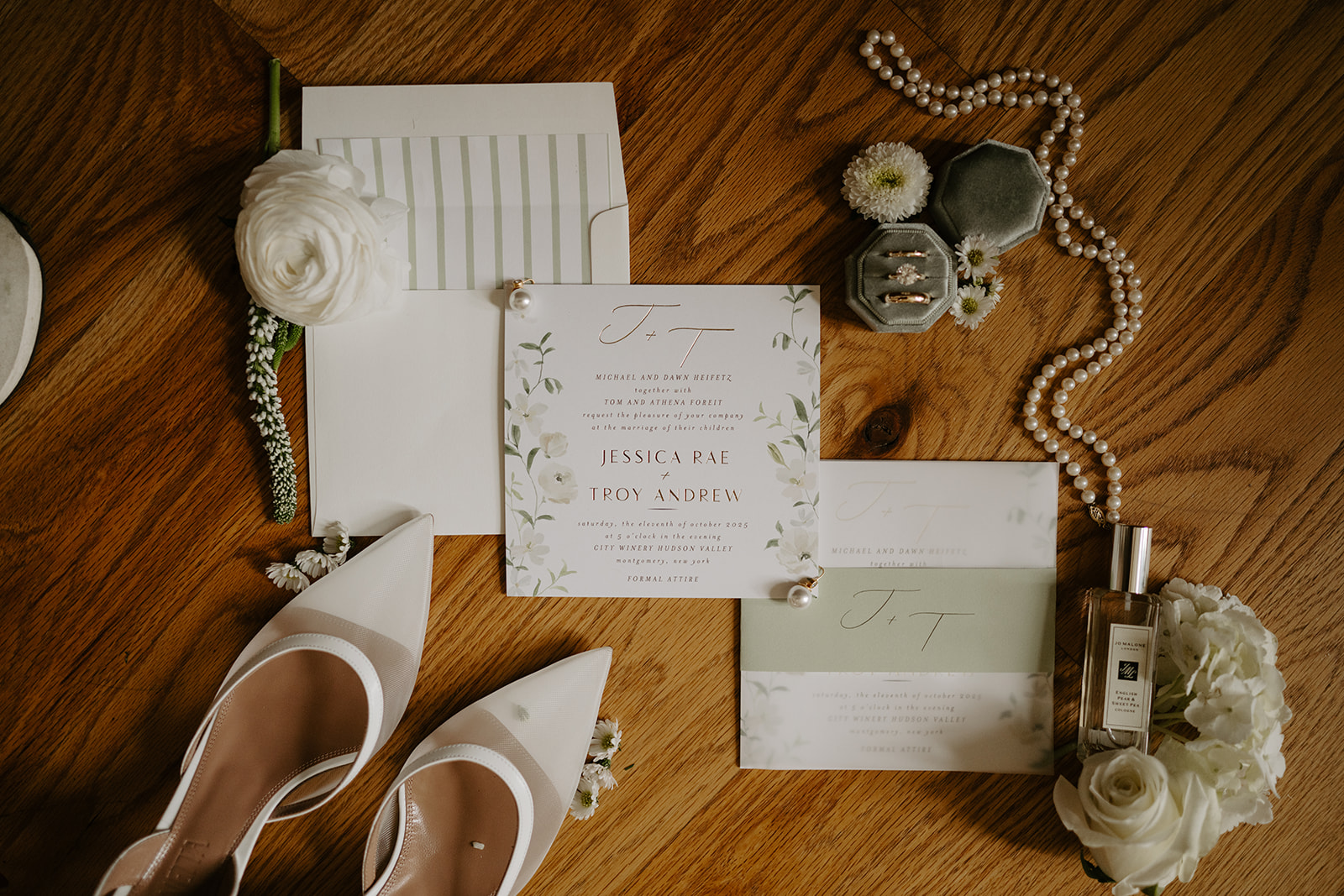 Flat lay of elegant wedding invitation suite, bridal shoes, rings, and pearl necklace on a wood floor.