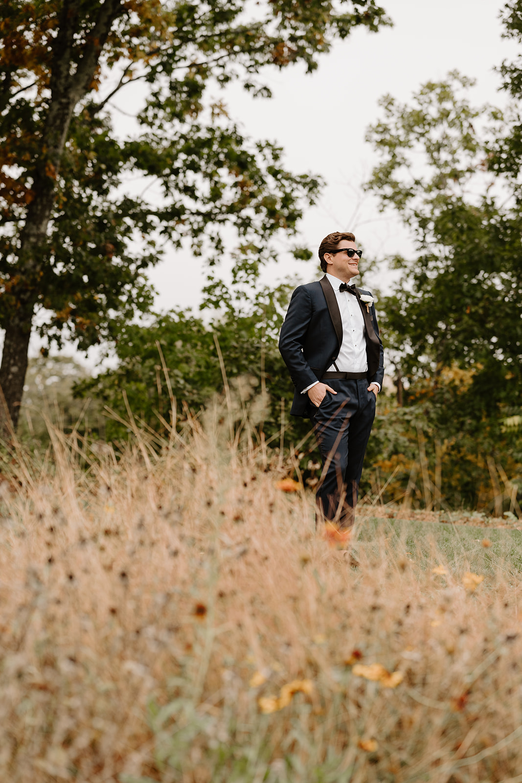 Groom in a dark tuxedo and sunglasses poses in a field of tall autumn grass, smiling.