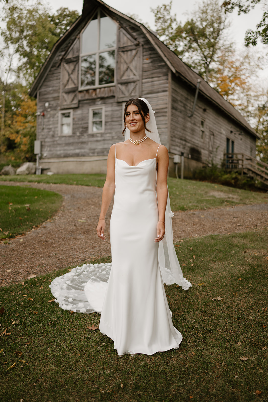 Bride in a white gown and veil stands smiling on the lawn in front of a rustic wooden barn.