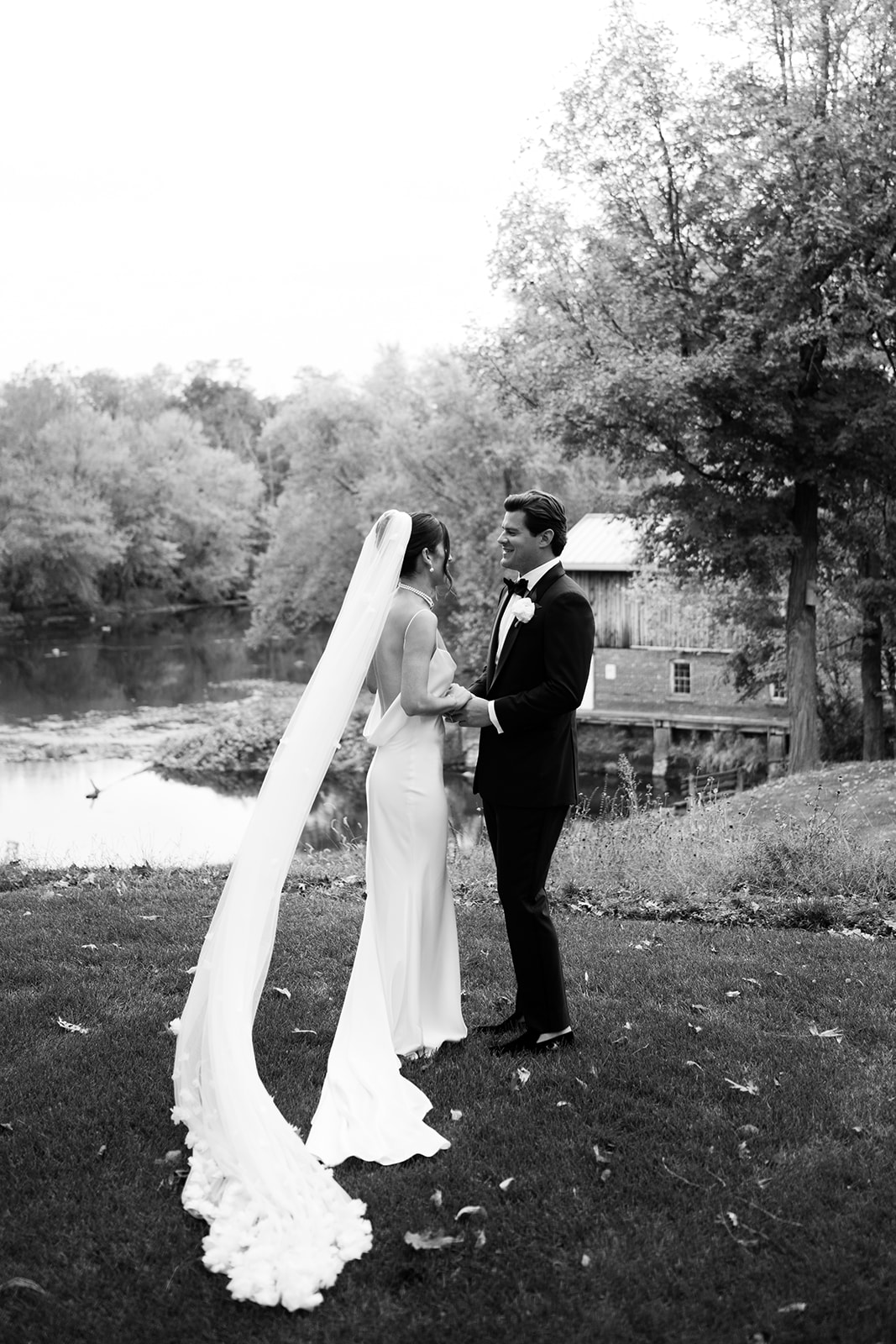 Black and white photo of the couple smiling at each other on a riverbank with a brick mill behind them.