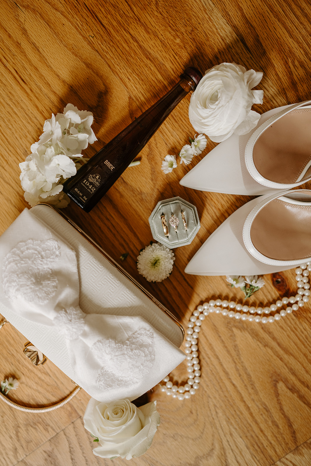 Flat lay of bridal accessories, featuring white heels, a custom tequila bottle, rings, and pearls.