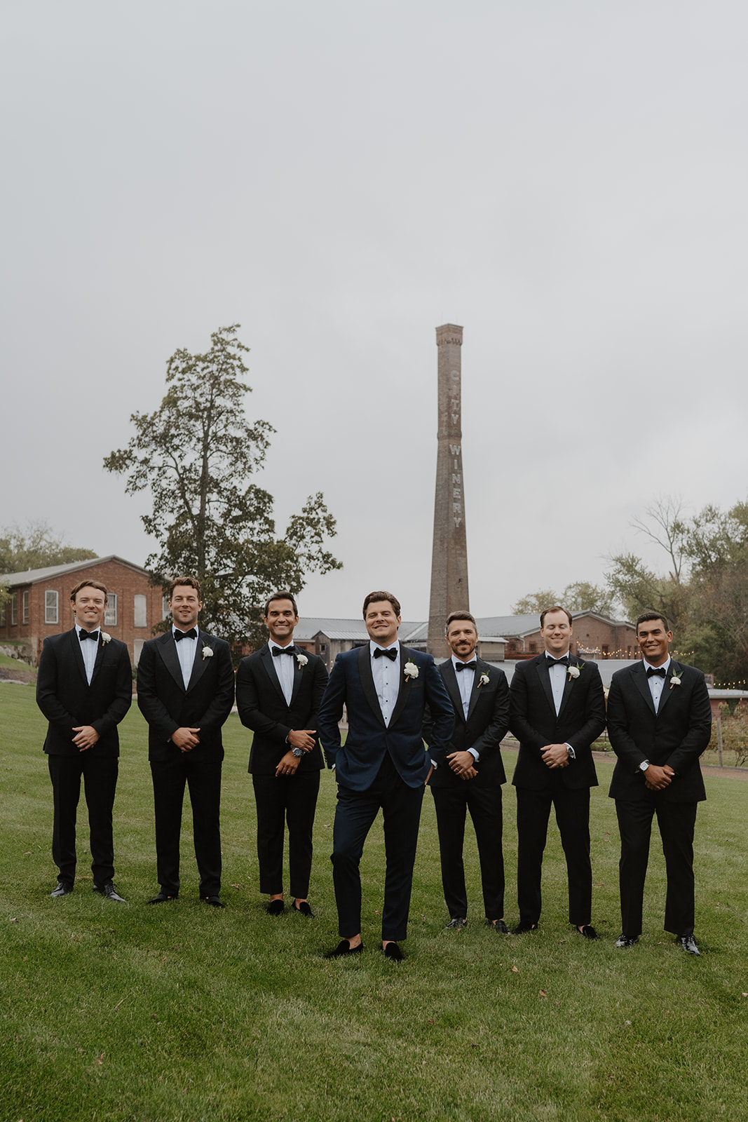 Groom in navy and groomsmen in black tuxedos stand on the green lawn at City Winery.