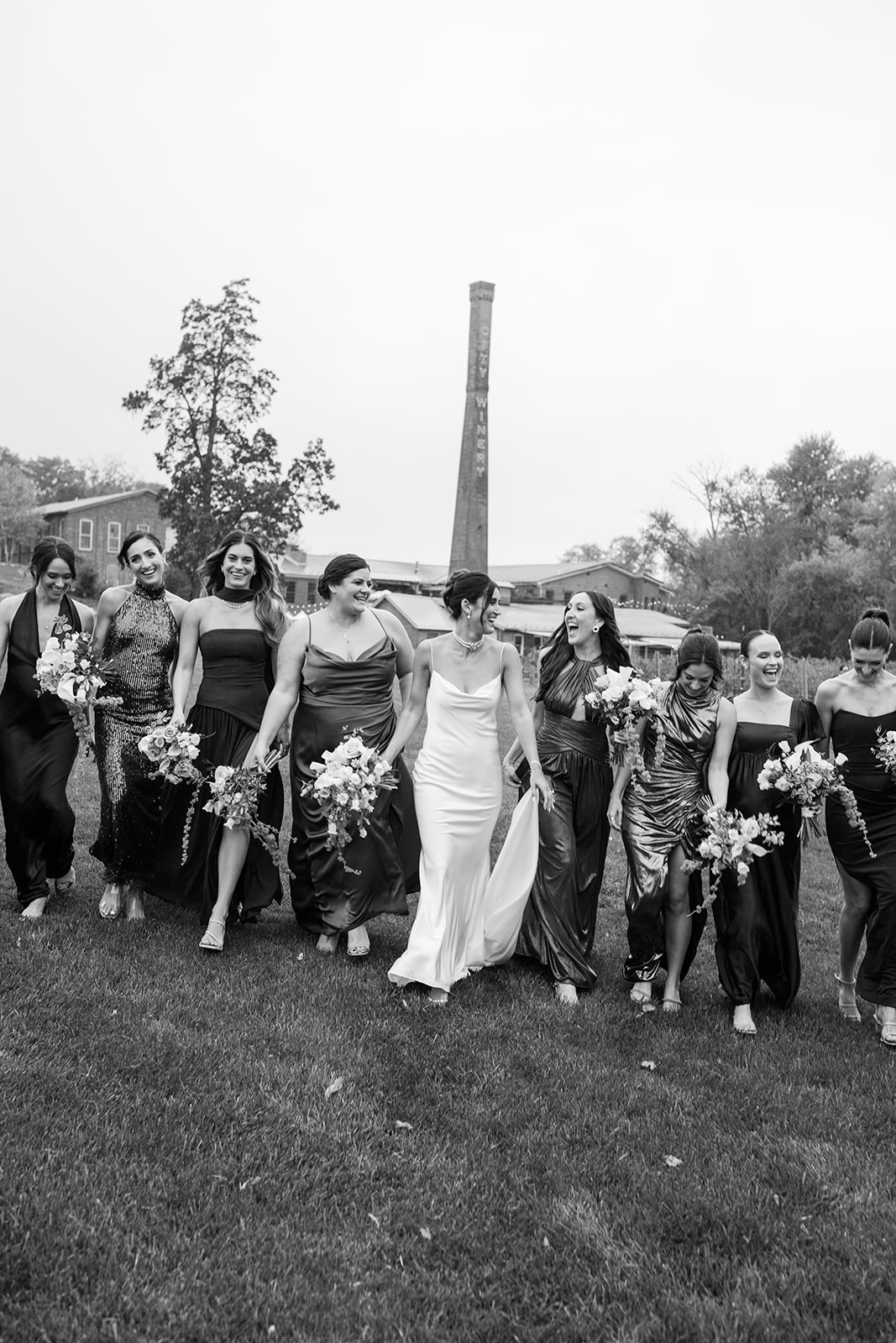Black and white photo of the bride and bridesmaids walking arm-in-arm, laughing and holding bouquets.
