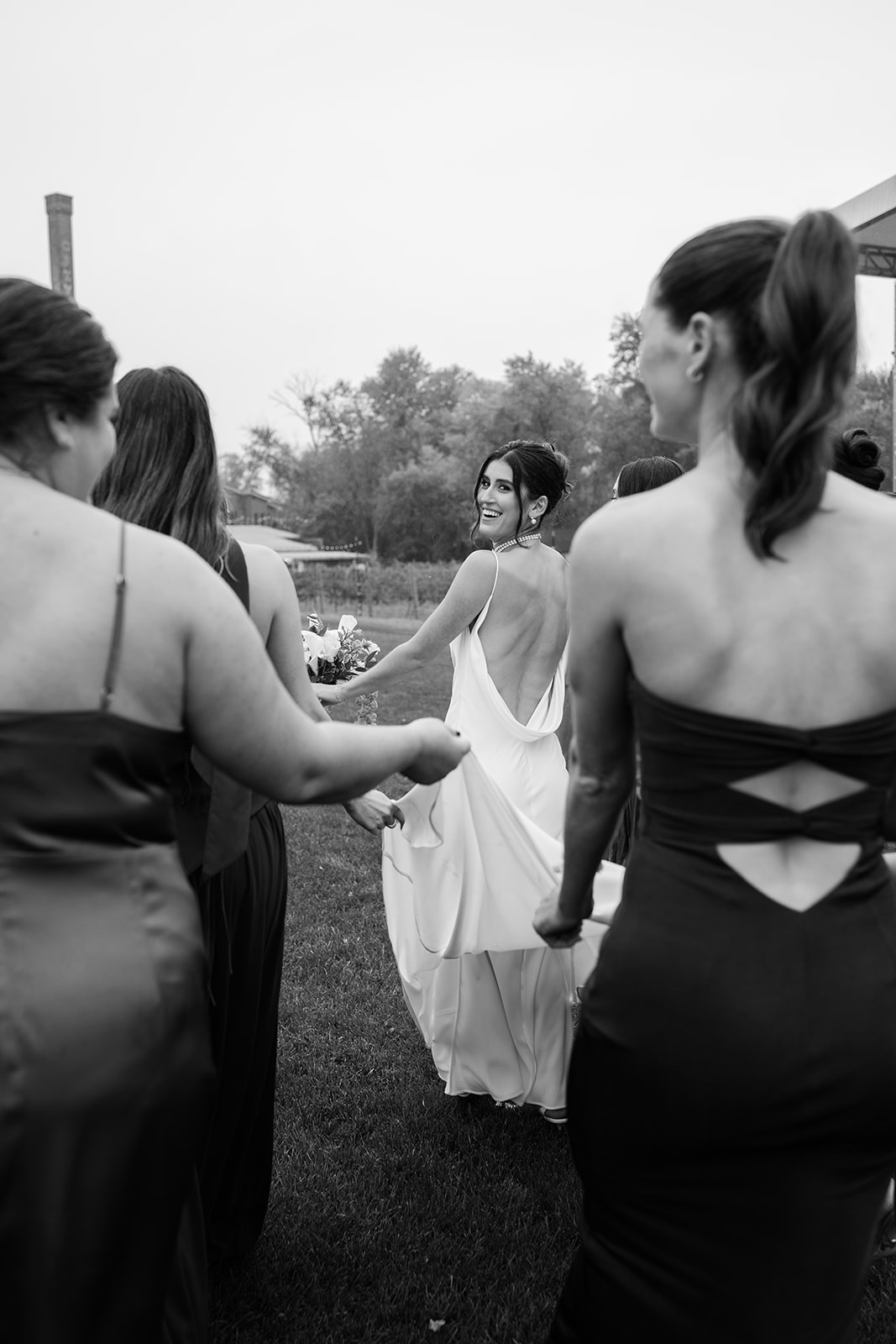 Black and white portrait of the bride looking back over her shoulder as bridesmaids hold her dress train.