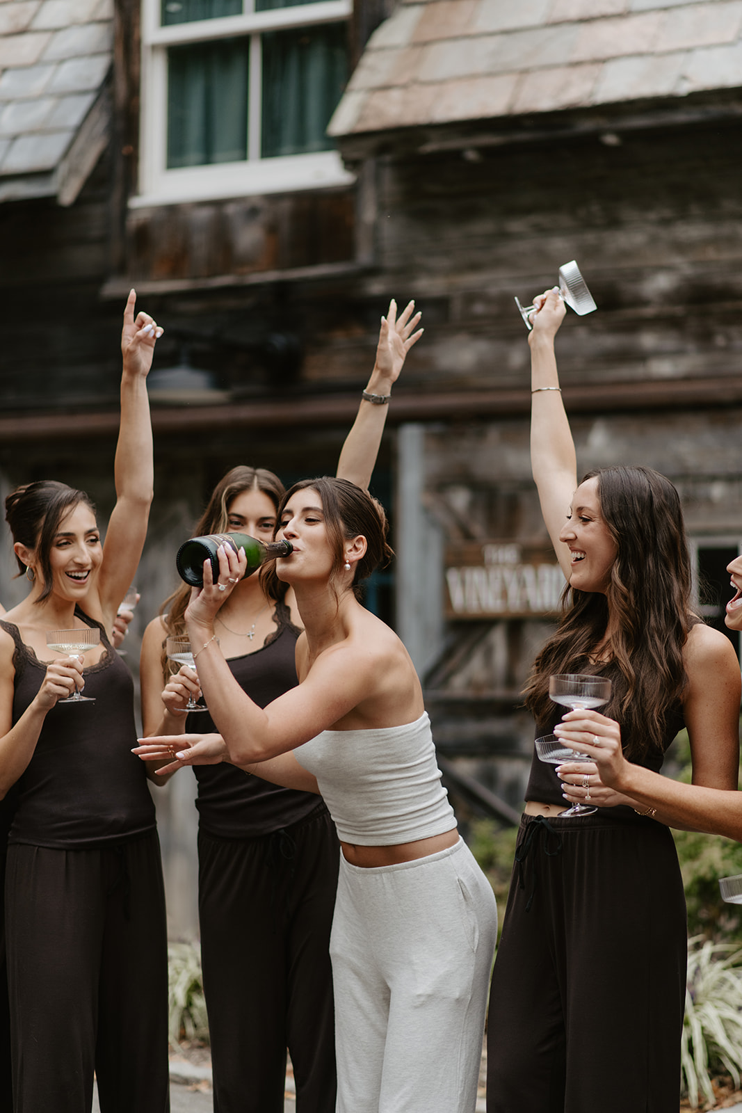 Bride drinks directly from a champagne bottle as bridesmaids cheer outdoors before the wedding.