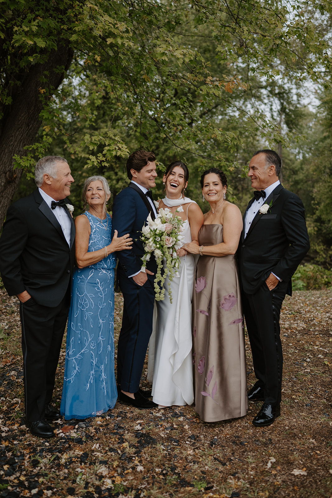 Bride and groom laugh heartily during outdoor portraits with their parents in the woods.