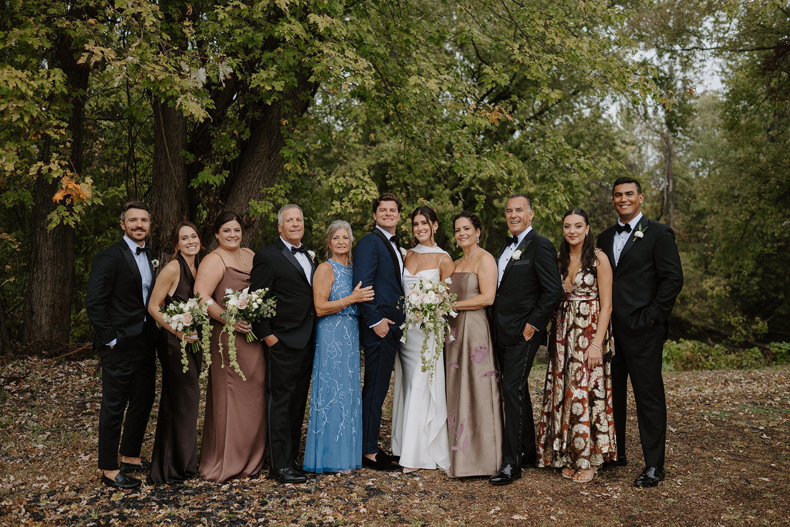 Extended family members smile together during outdoor group portraits in a wooded area.